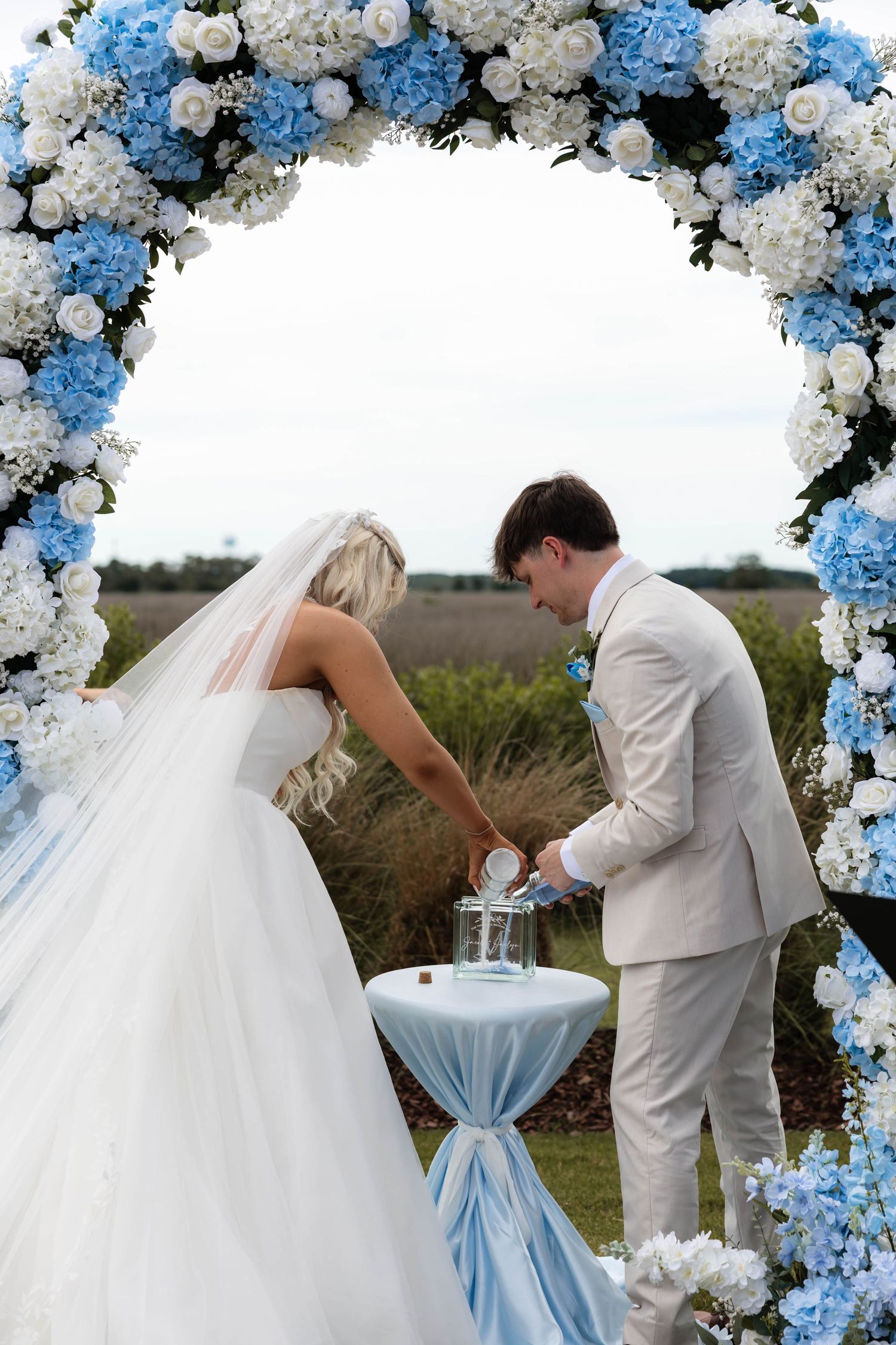 Bride and groom performing unity ceremony under blue and white floral arch at outdoor wedding.