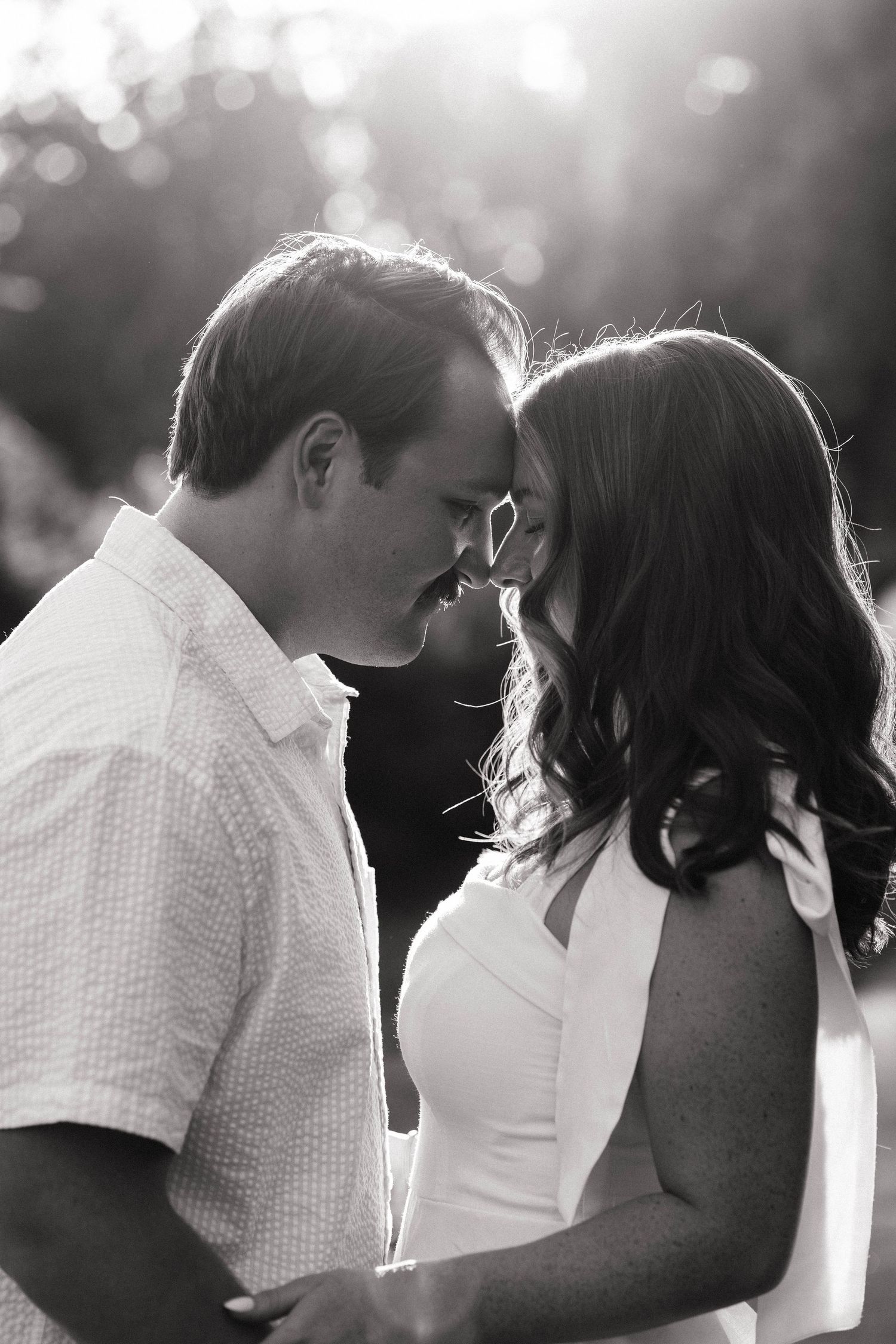 Black and white photo of a romantic couple leaning foreheads together, bathed in soft backlight outdoors.