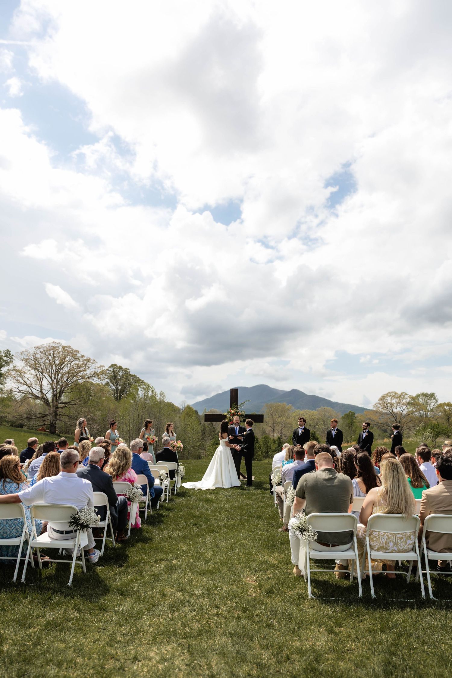 Outdoor wedding ceremony on green lawn with guests in white chairs, large cross, and mountain backdrop under cloudy sky.