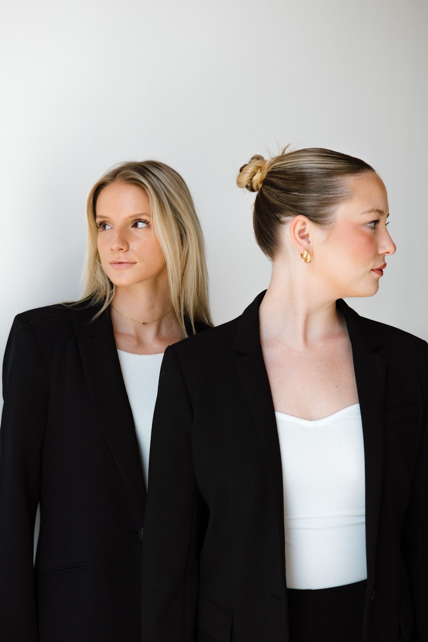 Two blonde women in black blazers and white tops pose back-to-back against a light gray background.