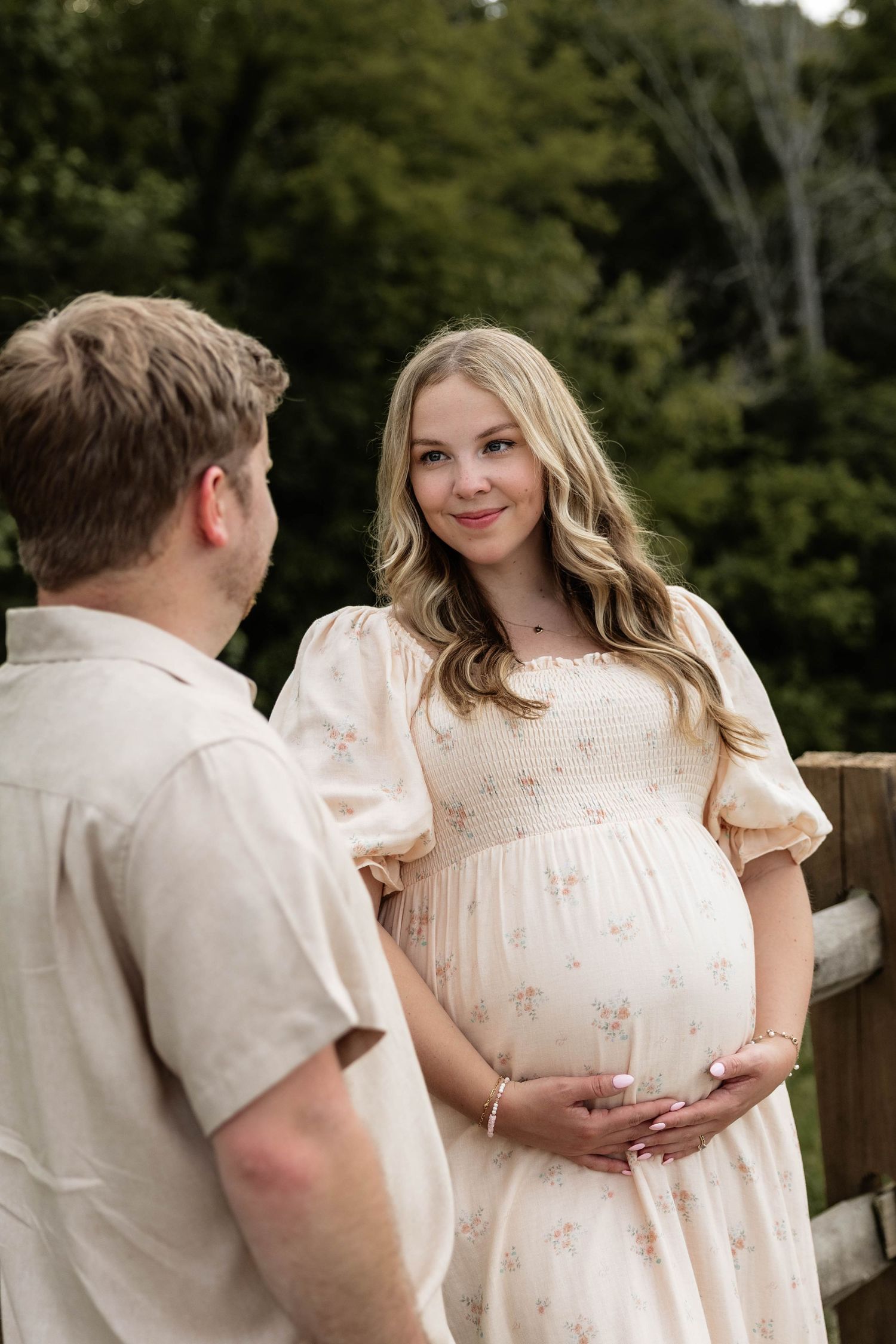 Pregnant woman in floral dress cradling baby bump while smiling at partner outdoors near wooden fence.