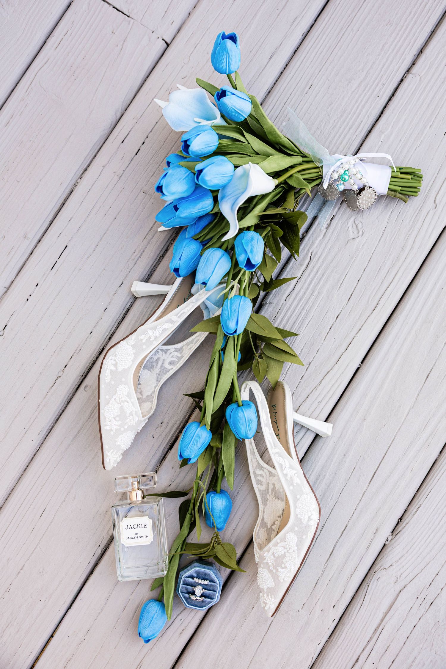 Blue tulip bridal bouquet with lace heels, perfume bottle, and ring box on rustic wooden floor.