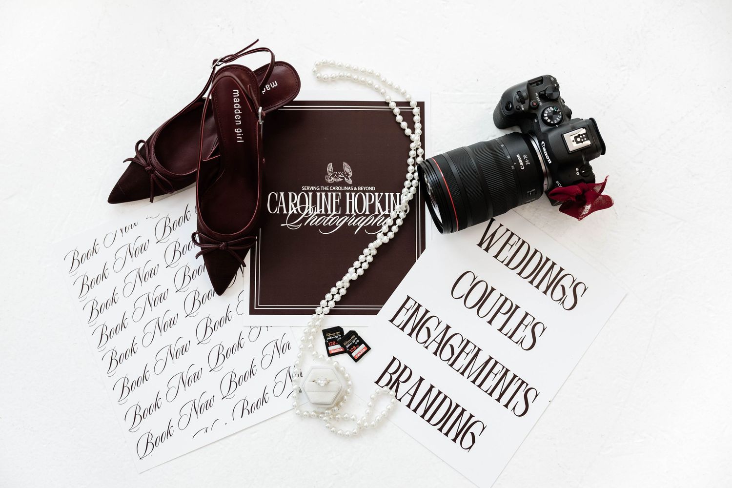Flat lay of Caroline Hopkins Photography branding with camera, pearl necklace, burgundy heels, and calligraphy cards on white background.