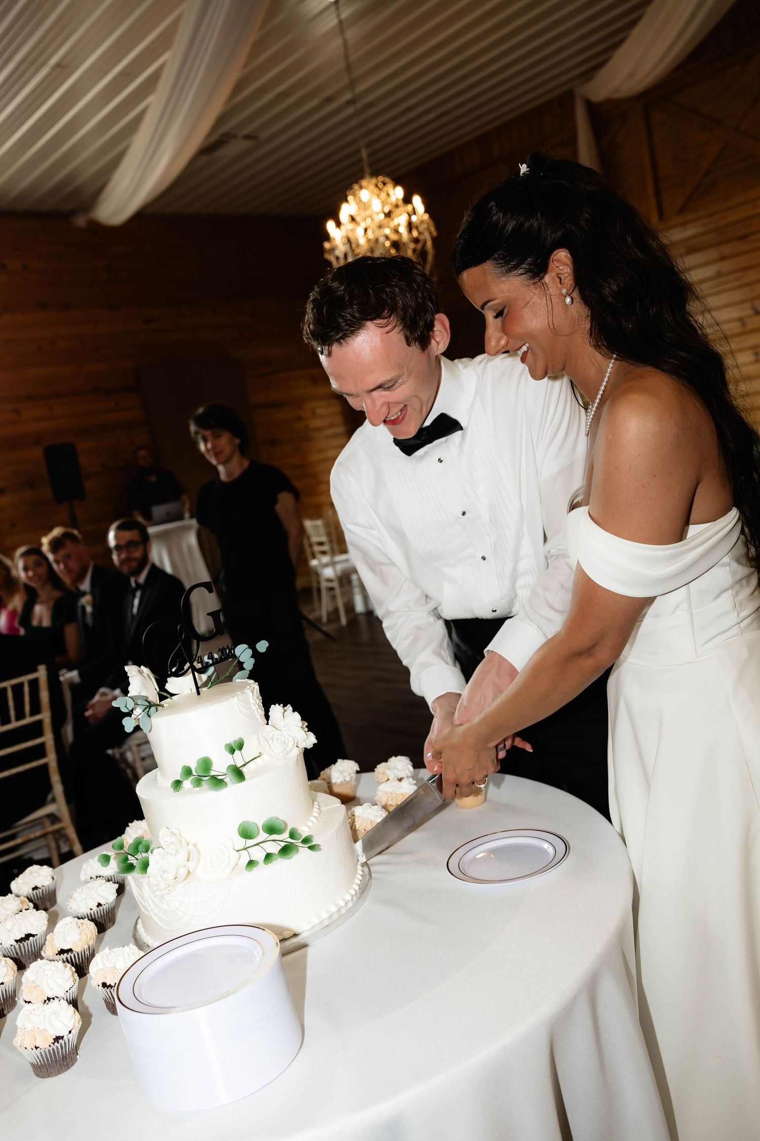 Bride and groom cutting white three-tier wedding cake decorated with greenery at elegant indoor reception venue.
