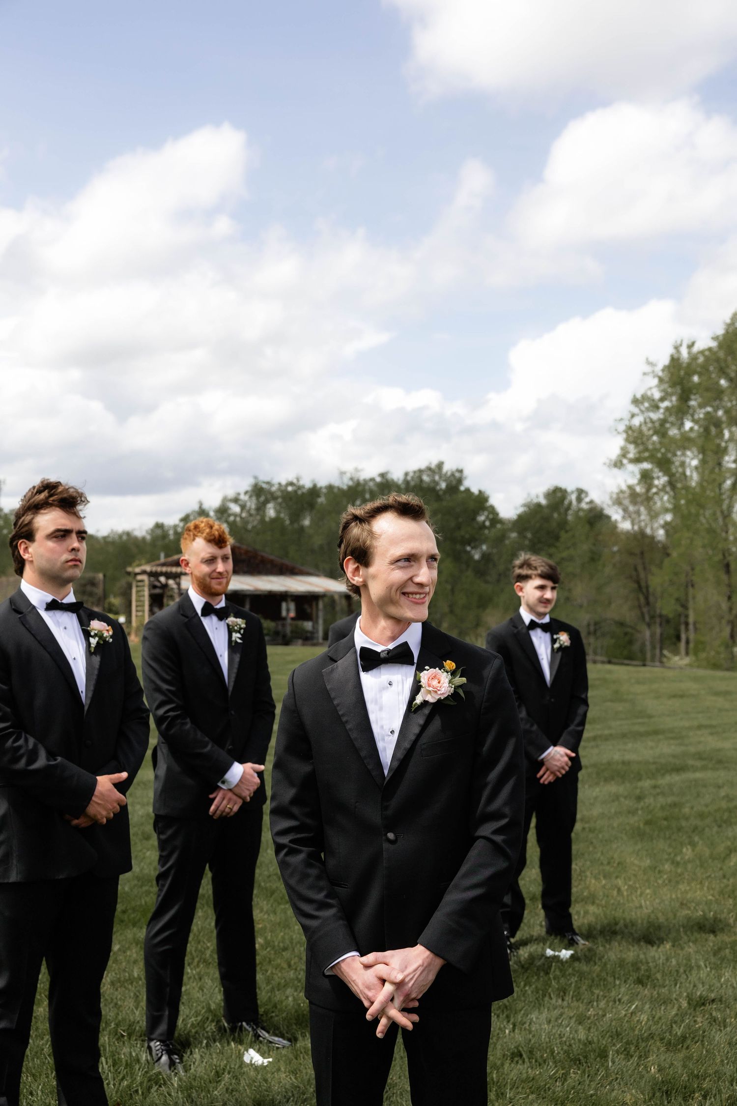 Groom in black tuxedo with floral boutonniere smiles outdoors, groomsmen standing behind on green lawn.