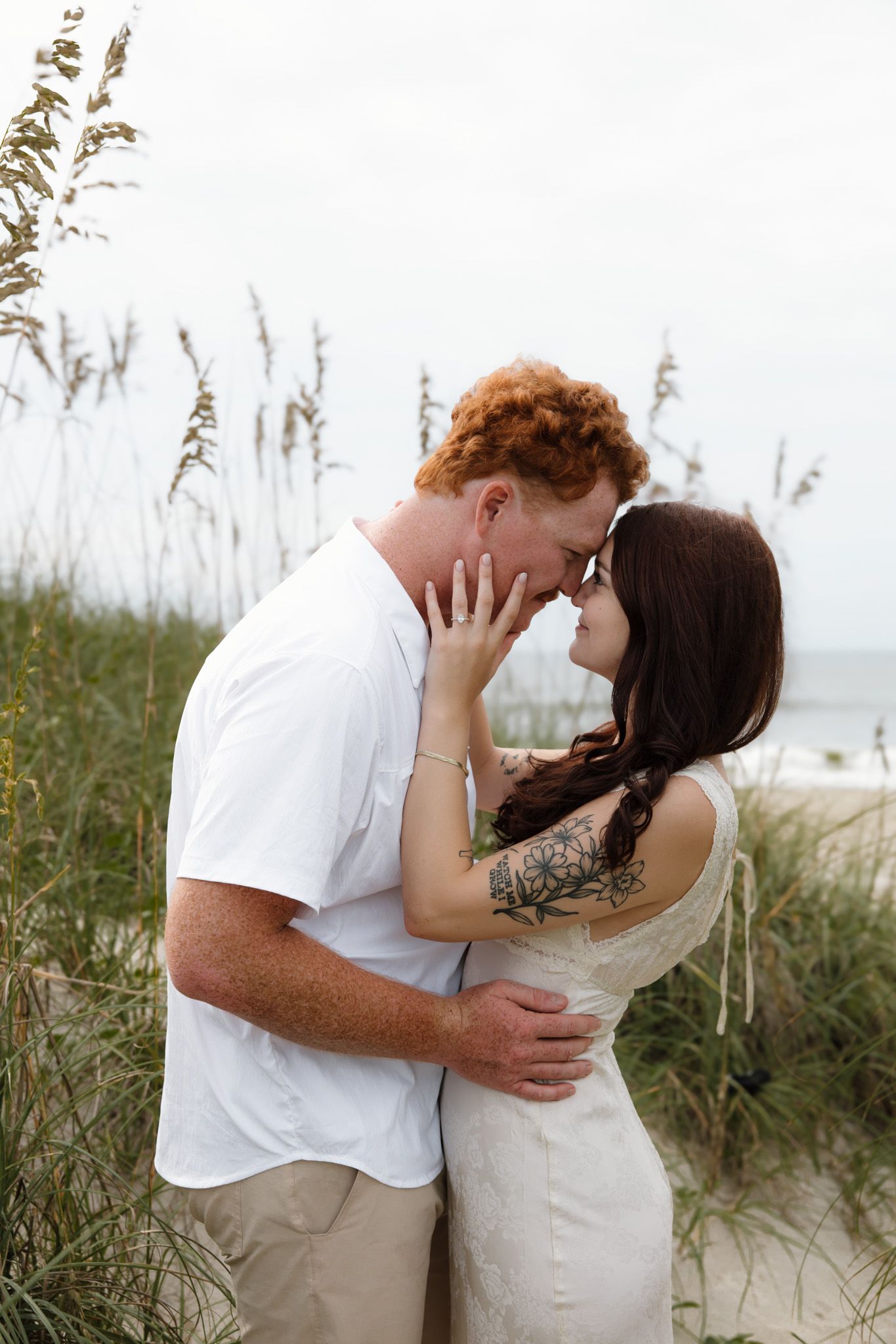 Romantic couple sharing an intimate moment among sea oats on a beach, woman in white dress with floral tattoo.