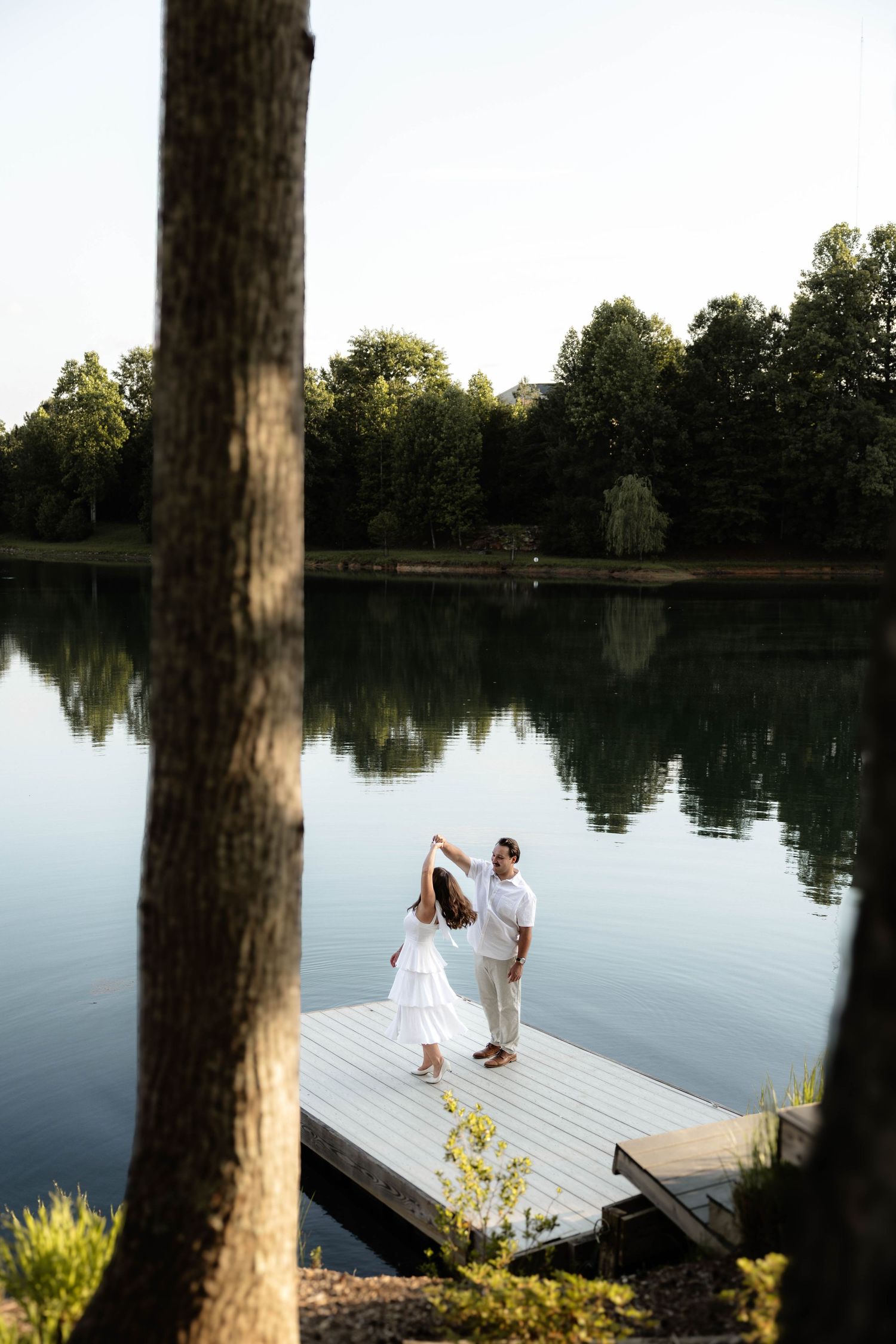 Couple dancing on a wooden dock by a calm lake surrounded by lush green trees on a serene evening.