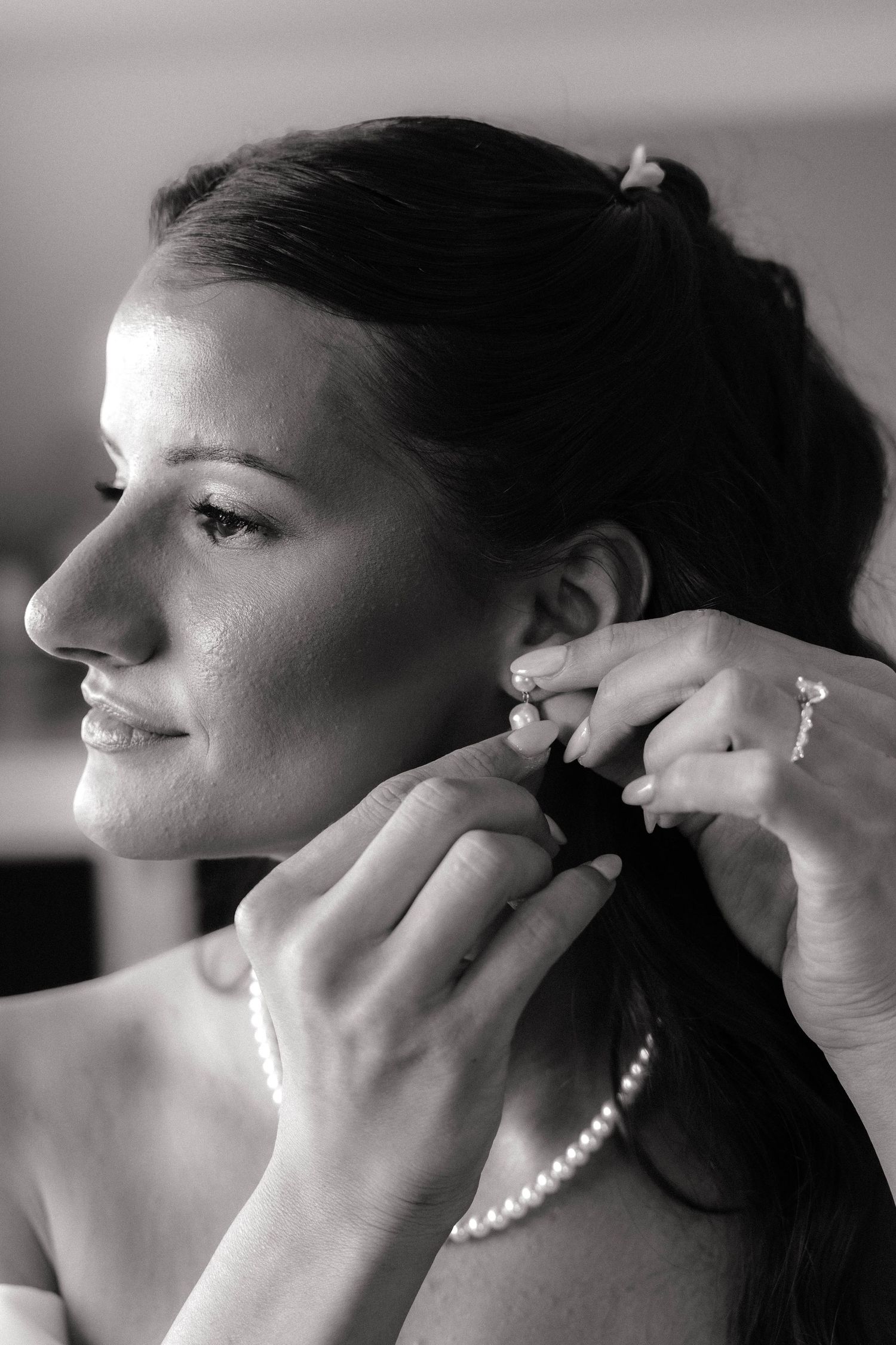 Black and white photo of a smiling bride having pearl earrings put on, wearing a pearl necklace.