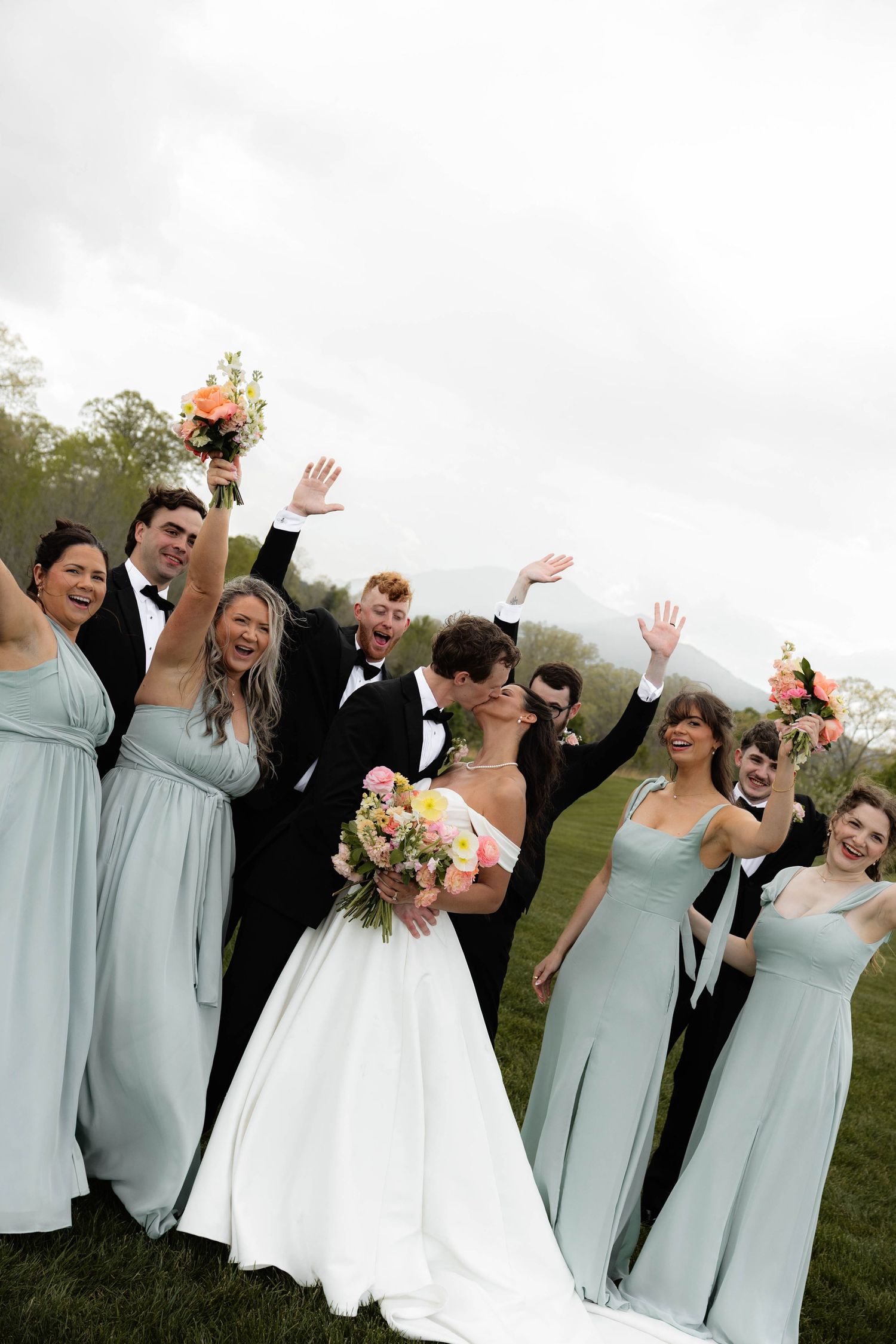 Wedding party celebrates as bride and groom kiss, bridesmaids in sage green gowns and groomsmen in tuxedos cheering outdoors.