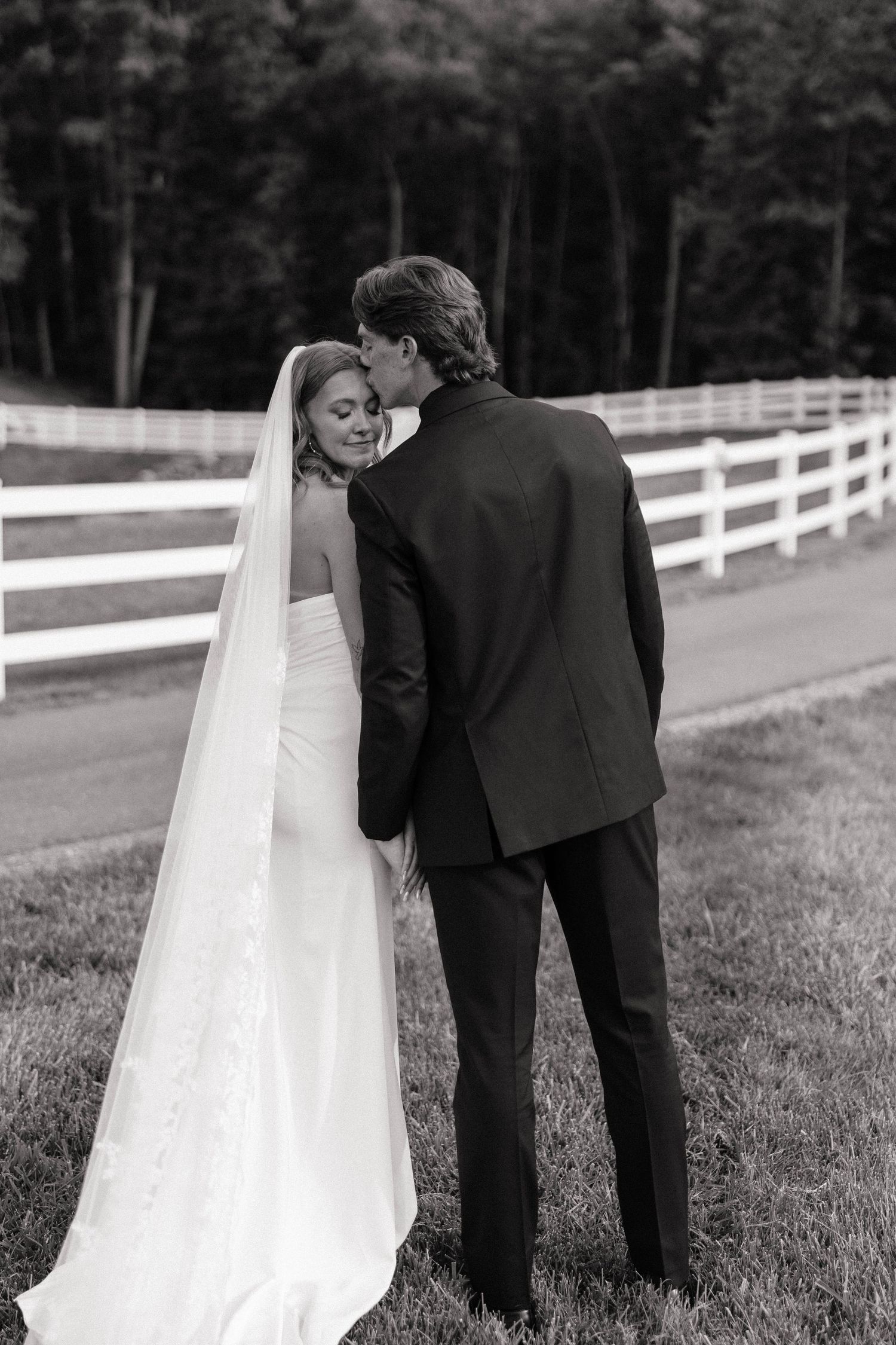 Black and white photo of a groom kissing his bride on the cheek outdoors near a white fence.