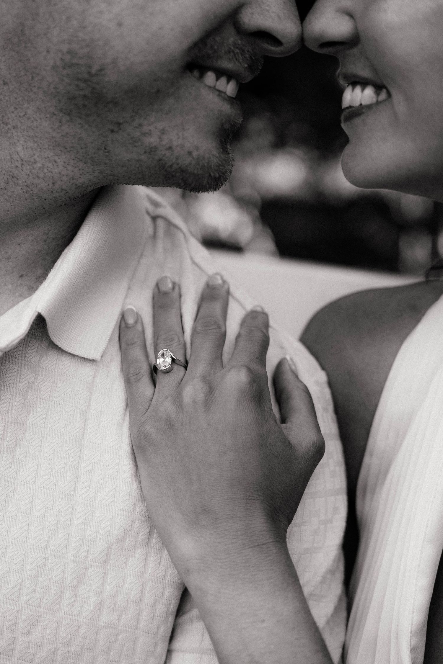 Black and white photo of smiling couple almost kissing, woman showing off diamond engagement ring on mans chest.