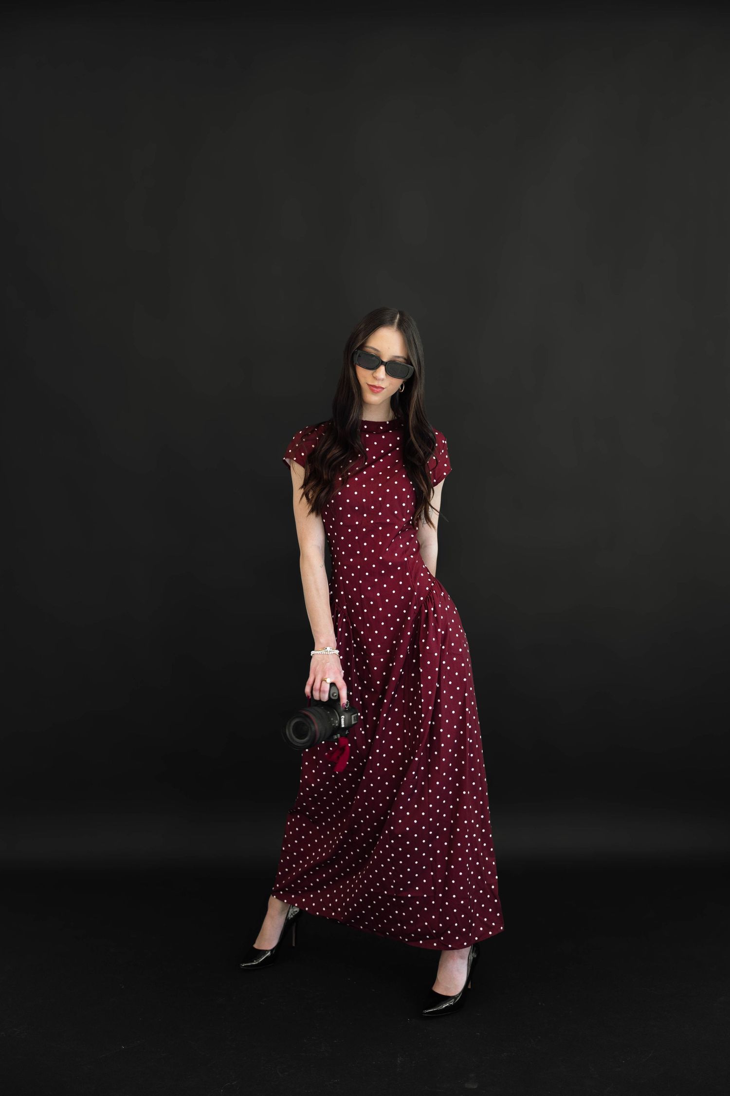 Woman in red polka dot maxi dress with sunglasses and black heels posing against dark background.