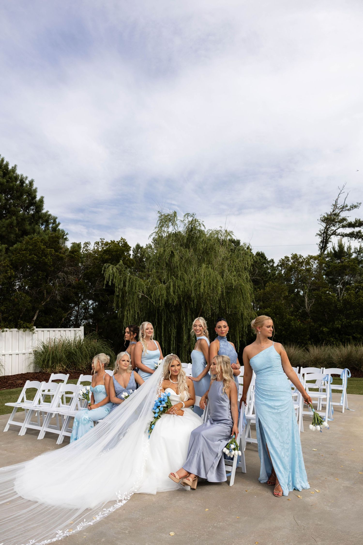 Bride in white gown with long veil sits among bridesmaids in light blue dresses at outdoor wedding ceremony.