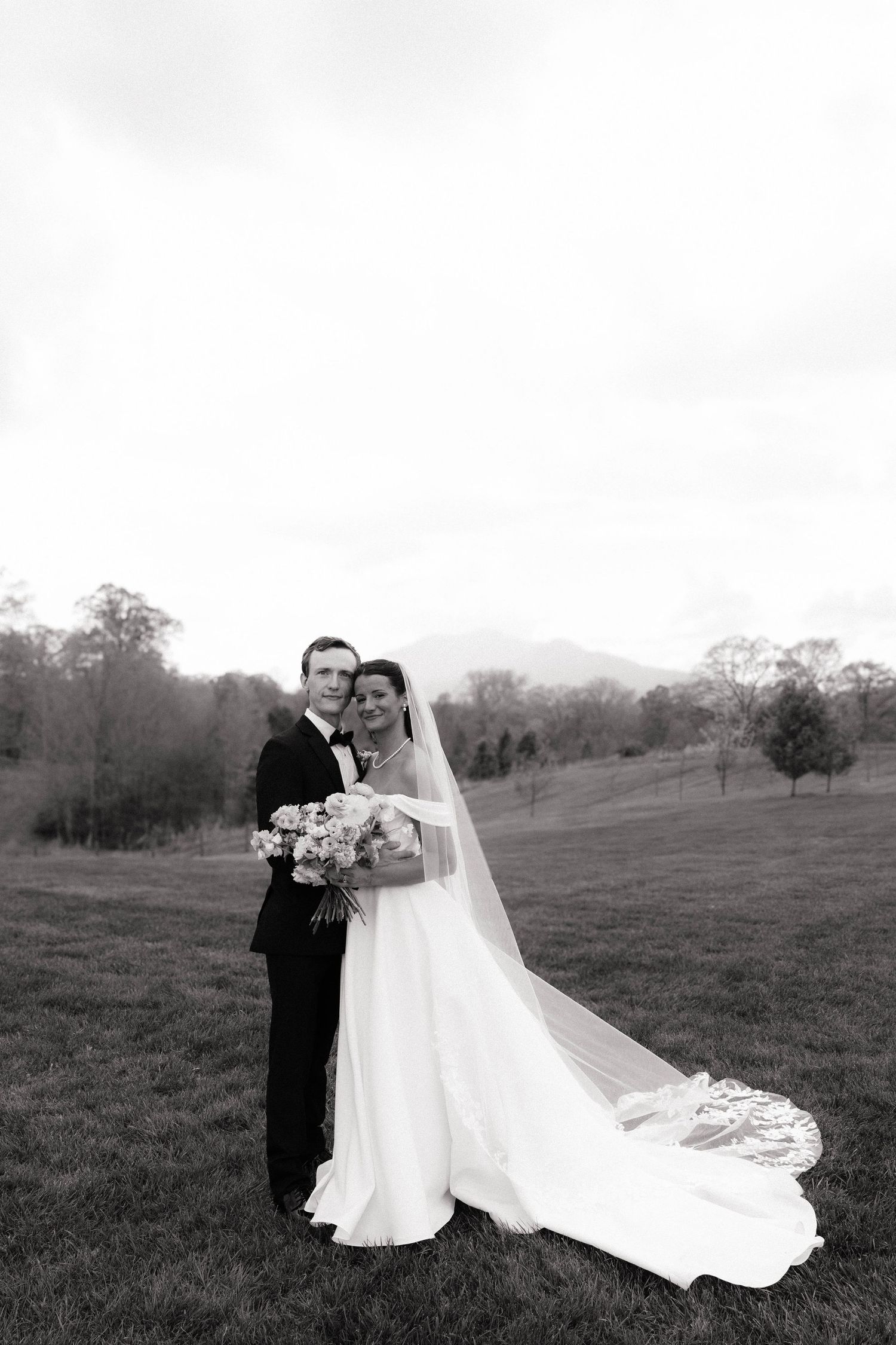Black and white photo of a bride and groom posing outdoors on a grassy field with mountains in the background.