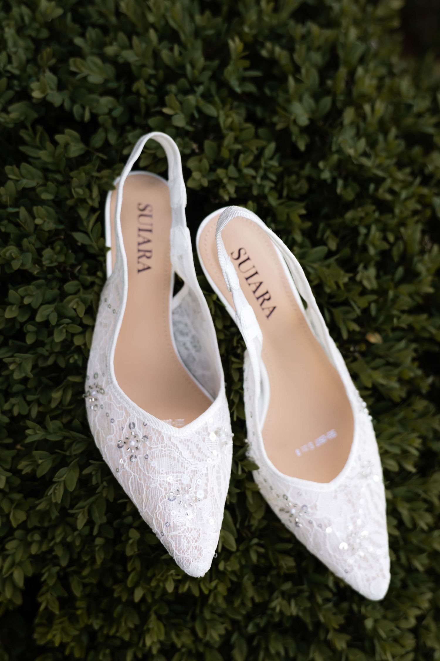 White lace slingback bridal flats with crystal embellishments by Suara, displayed on green boxwood hedge.