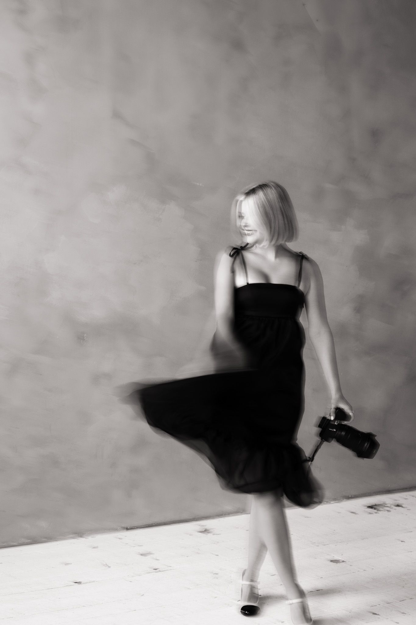 Blond woman in black dress holding camera, captured in motion blur, black and white studio photography.