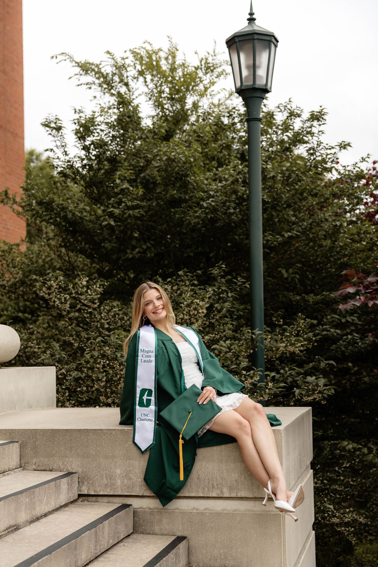 Young woman in green graduation cap and gown sitting on stone steps near a lamp post, smiling at her graduation.