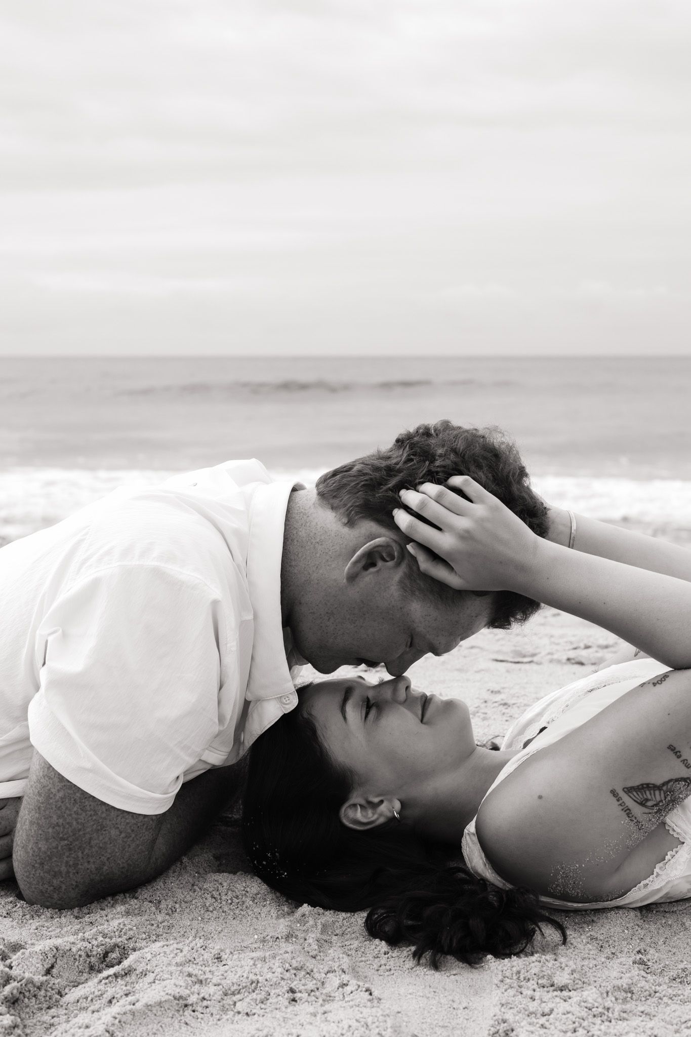 Black and white photo of a romantic couple sharing a tender moment on a sandy beach with ocean waves in the background.