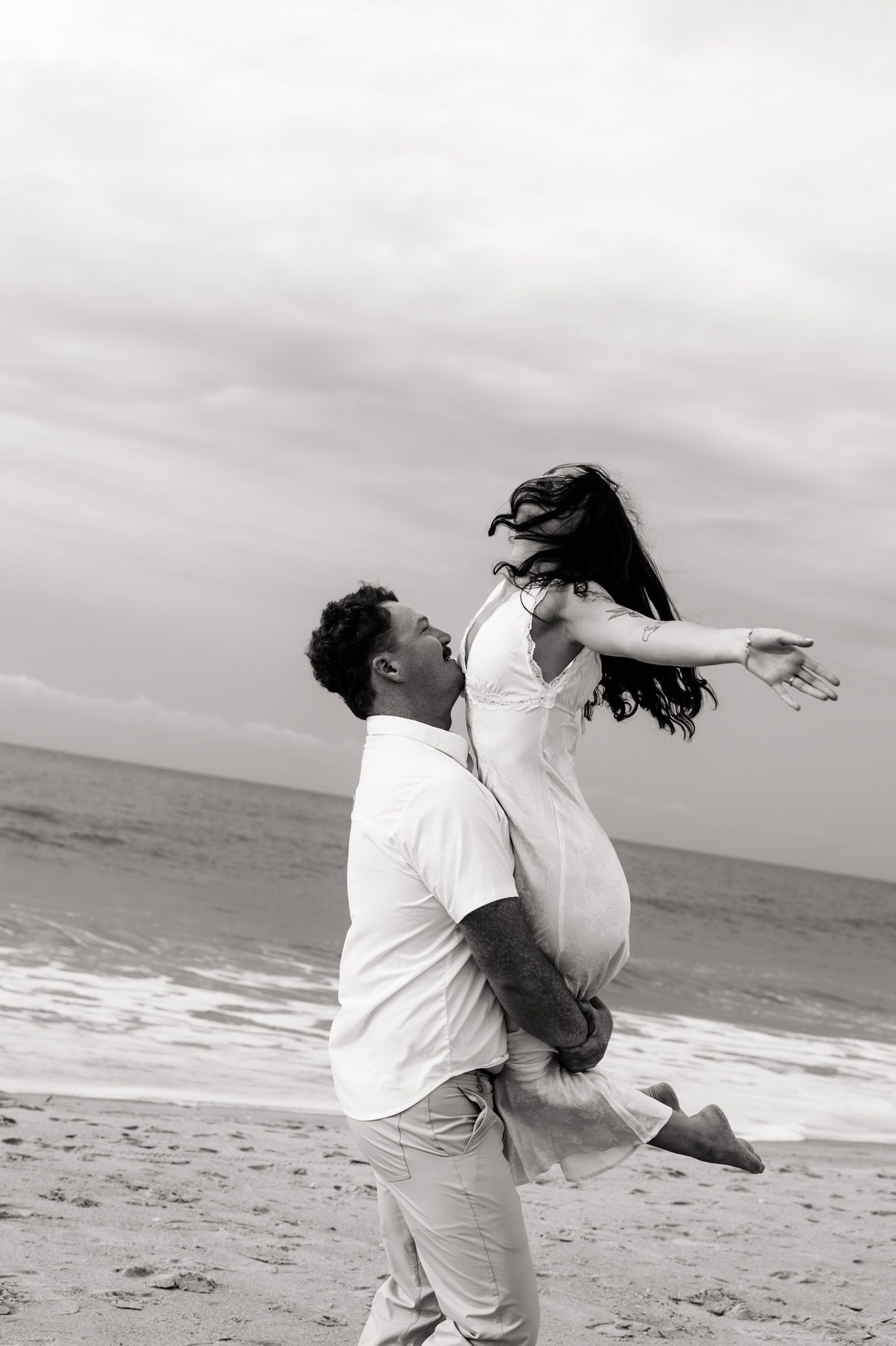 Black and white photo of a man lifting a woman in a white dress on a beach, with ocean waves in the background.