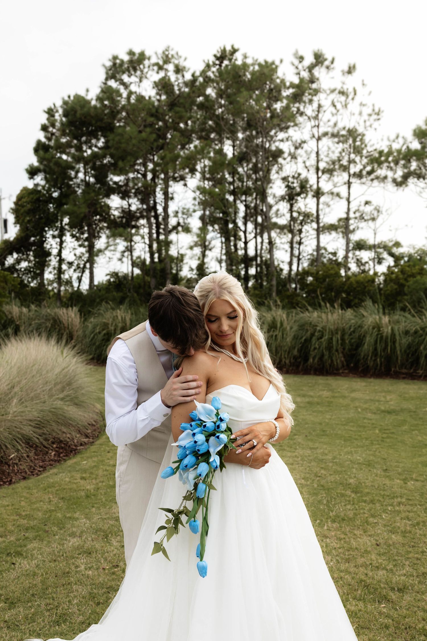 Bride in white gown holding blue bouquet embraced by groom in gray suit outdoors with tall trees and greenery.