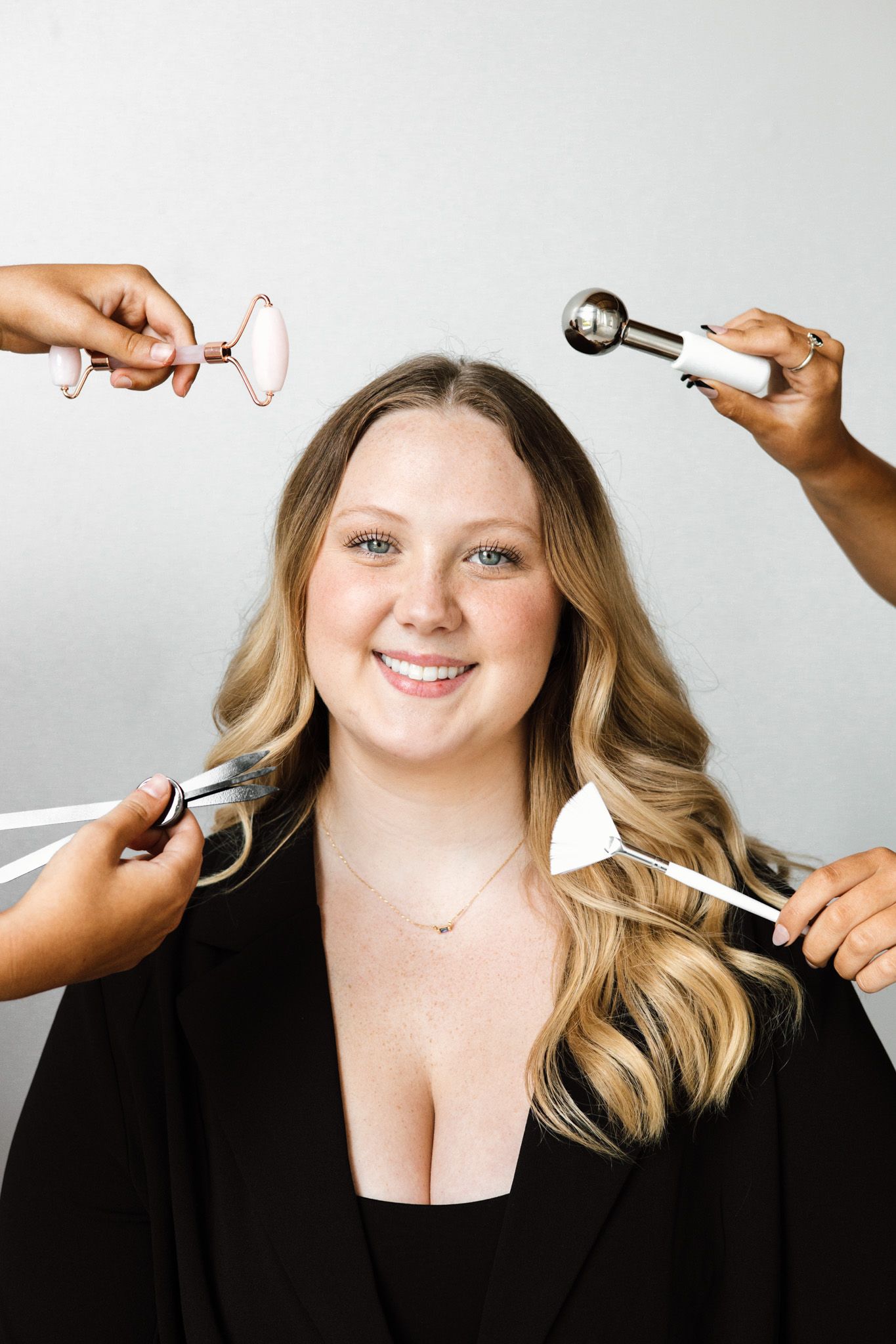 Smiling woman with wavy blonde hair in black blazer surrounded by hands holding beauty tools and skincare devices.