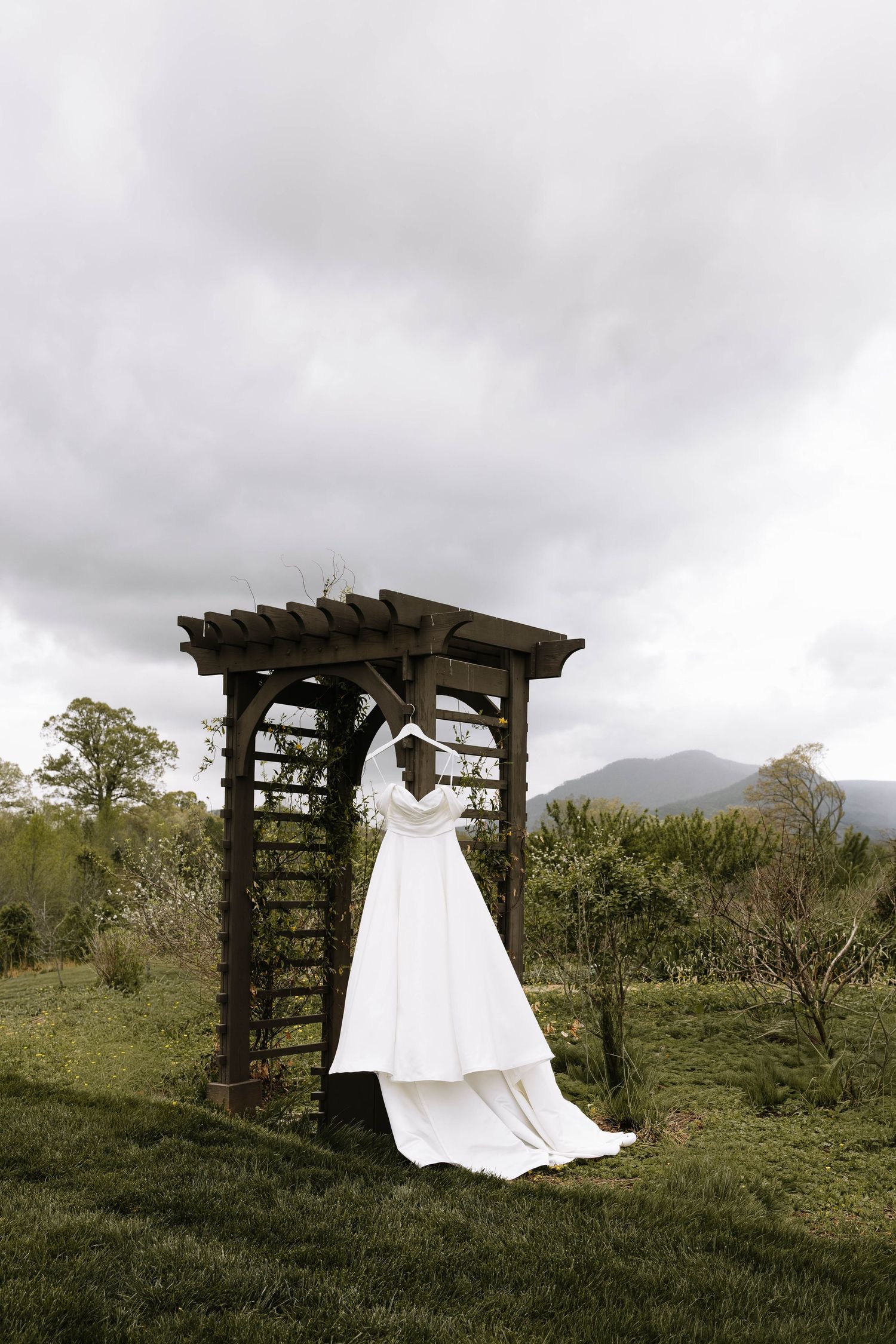 White wedding dress hanging on a rustic wooden arbor in a green meadow with misty mountains in the background.