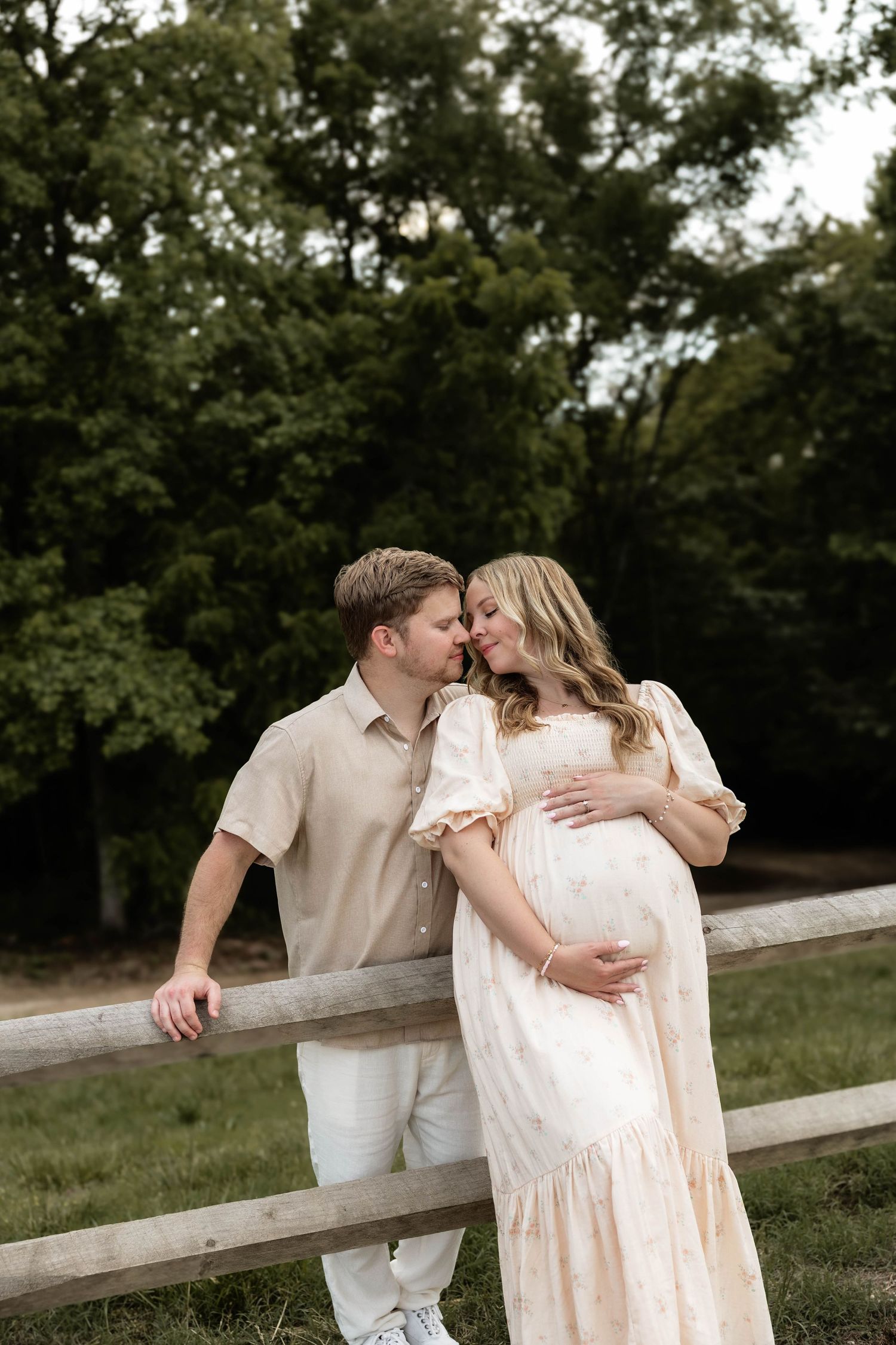 Couple sharing a tender moment by a wooden fence outdoors, pregnant woman in flowy peach dress cradling her baby bump.