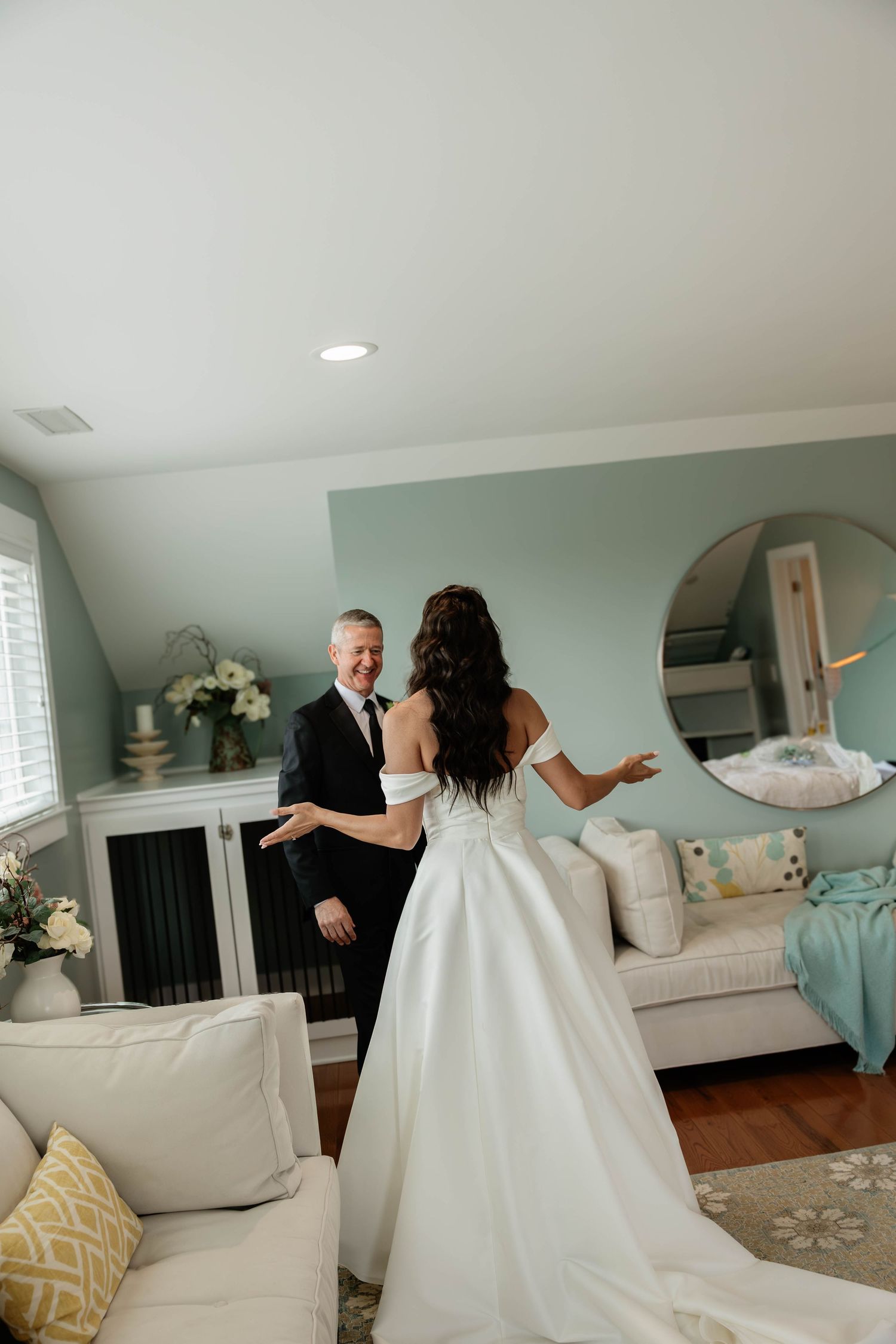 Bride in white gown shares first look moment with father in a light blue room, both smiling with arms outstretched.