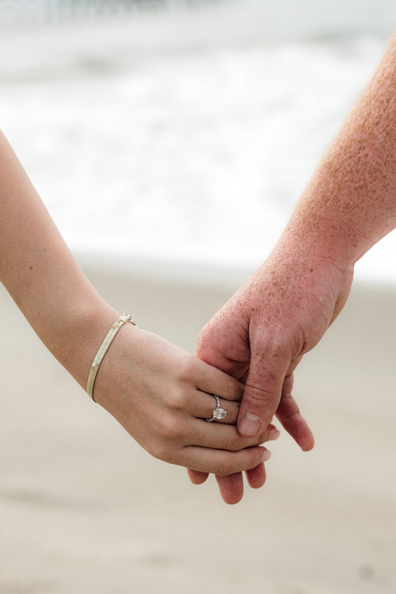 Couple holding hands on beach, woman wearing diamond engagement ring and gold bracelet, ocean waves in background.