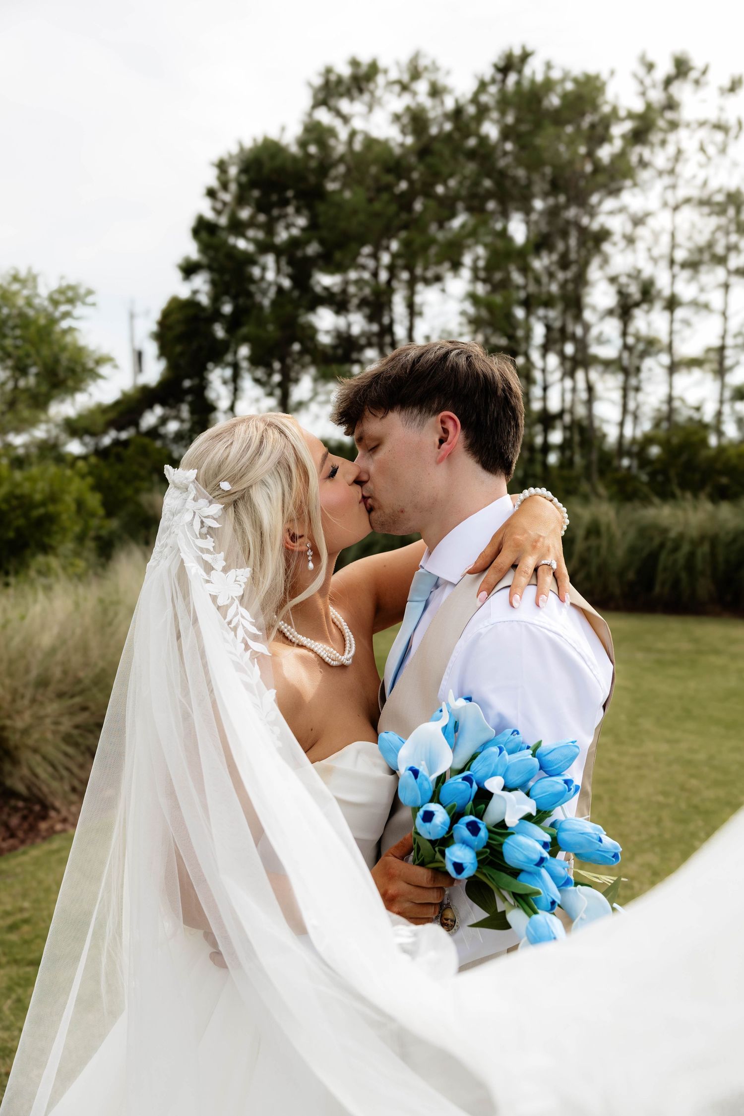 Bride and groom share a romantic kiss outdoors, bride holding blue tulip bouquet with flowing white veil.