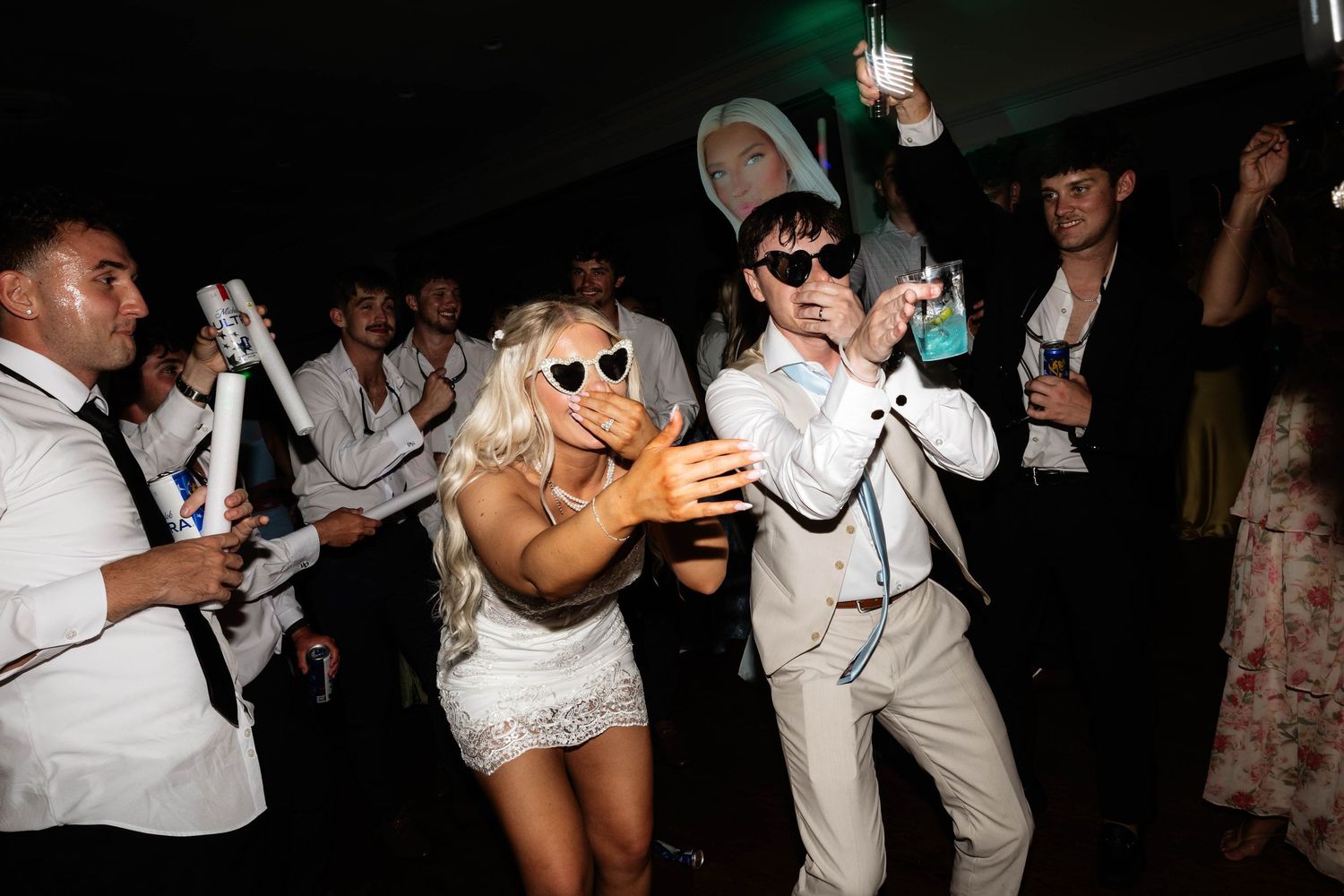 Bride and groom dancing at wedding reception party with guests holding glow sticks in dark venue.