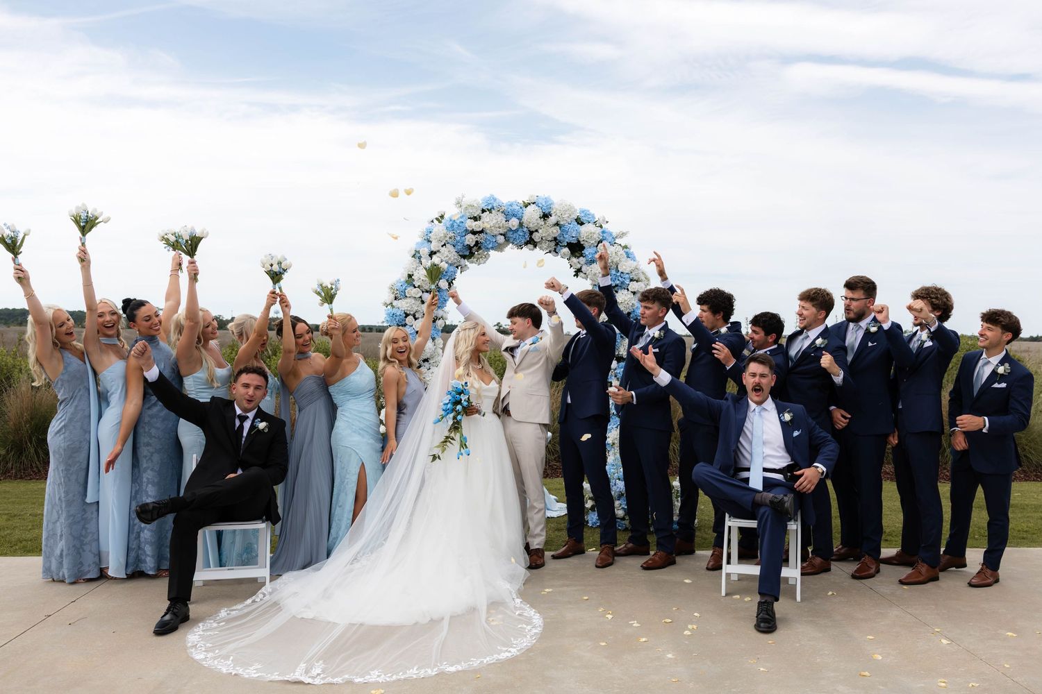 Wedding party celebrates outdoors under a blue floral arch, bridesmaids in blue gowns and groomsmen in navy suits.