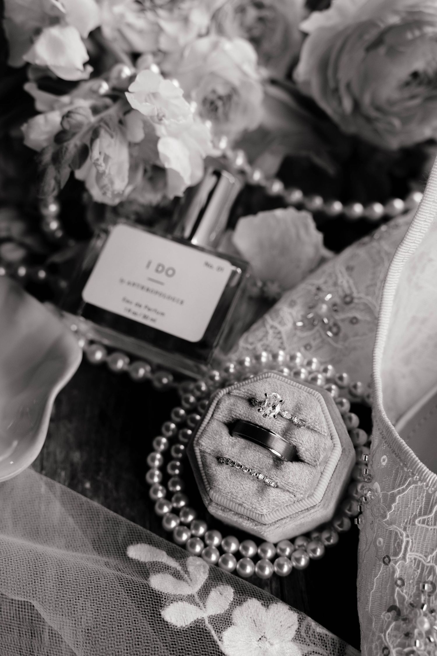 Black and white wedding flat lay with diamond rings in velvet box, pearl necklace, I Do perfume bottle, and floral bouquet.