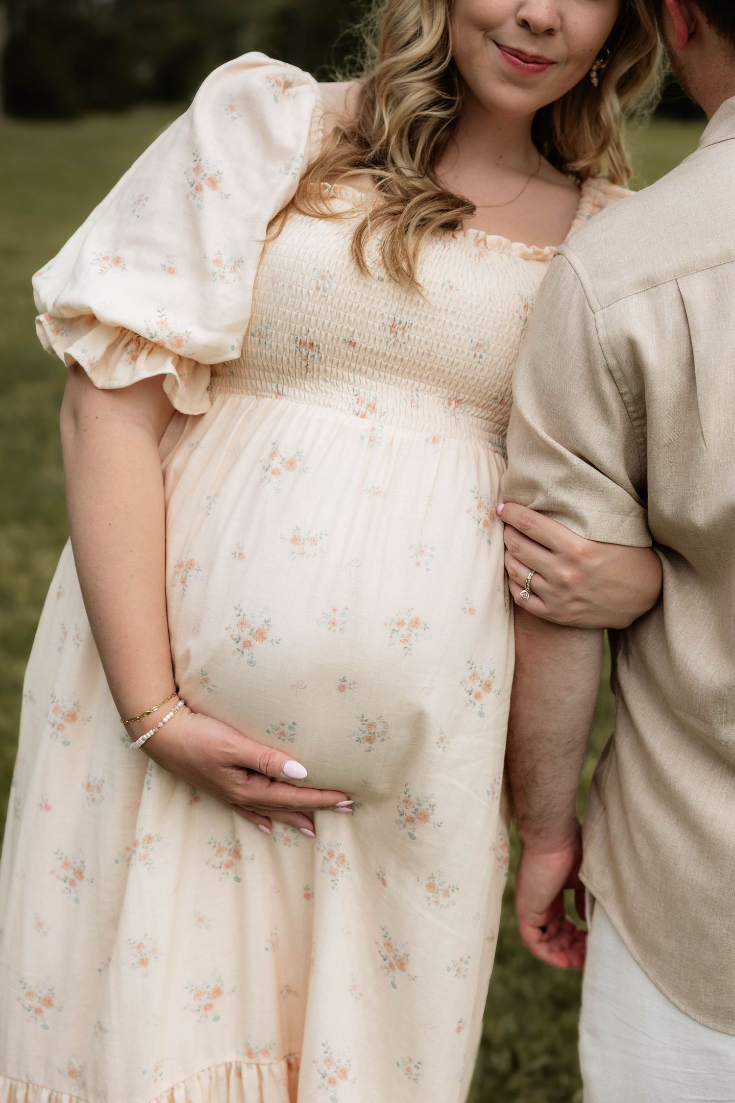 Pregnant woman in floral smocked dress holds baby bump outdoors while partner in linen shirt stands beside her.