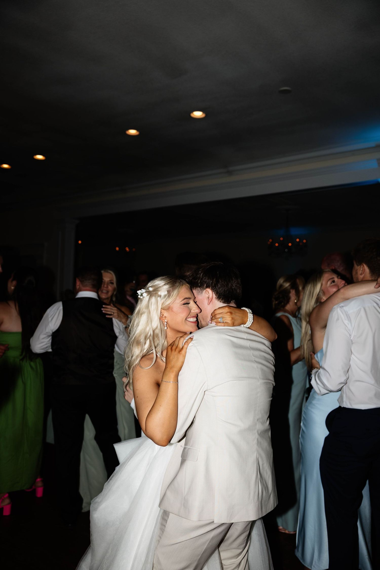 Bride and groom share a joyful embrace on the dance floor surrounded by wedding guests at their reception.