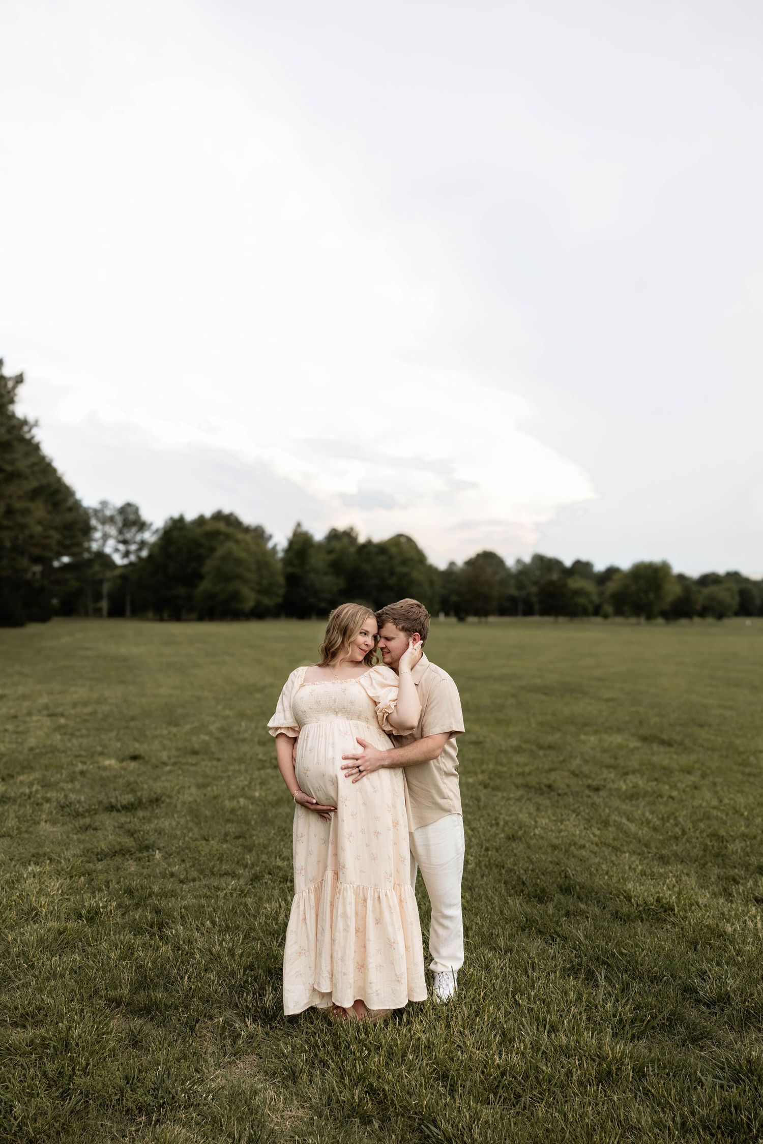 Couple sharing tender moment in green field, pregnant woman in pink lace dress embraced by partner in white outfit.