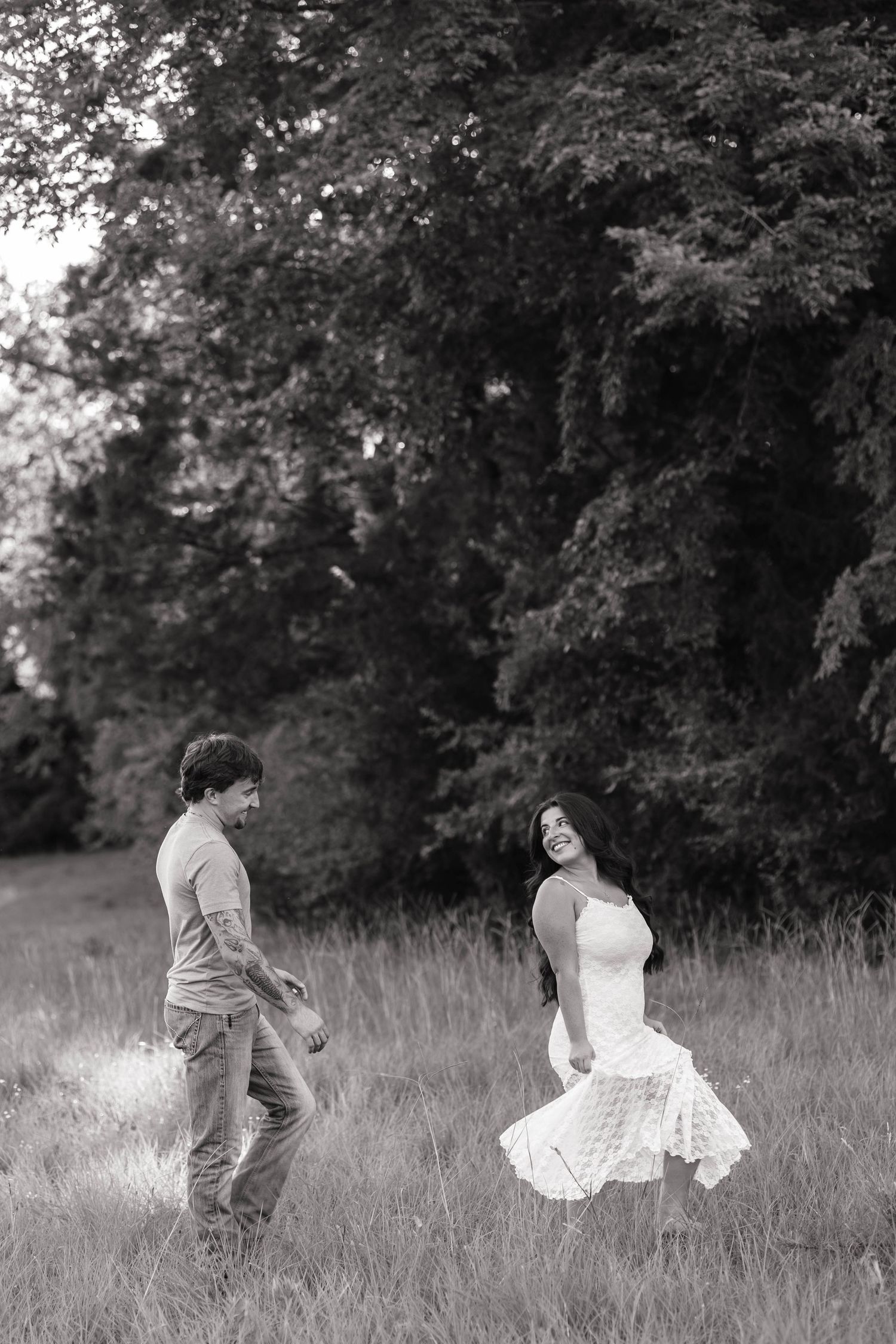 Black and white photo of a joyful couple dancing in a meadow surrounded by lush trees.