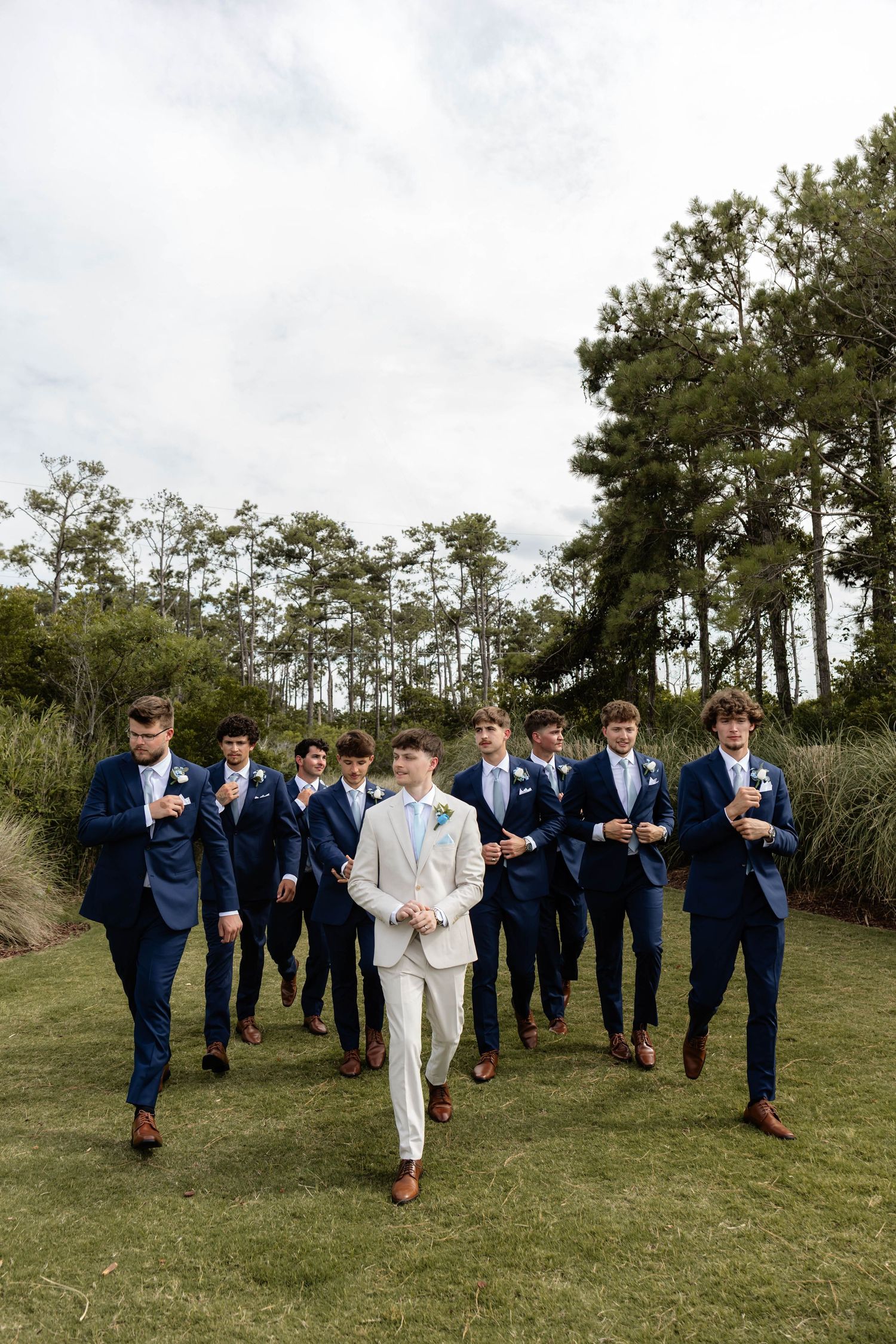 Groom in light gray suit leads groomsmen in navy blue suits walking outdoors on green lawn with tall pine trees behind.