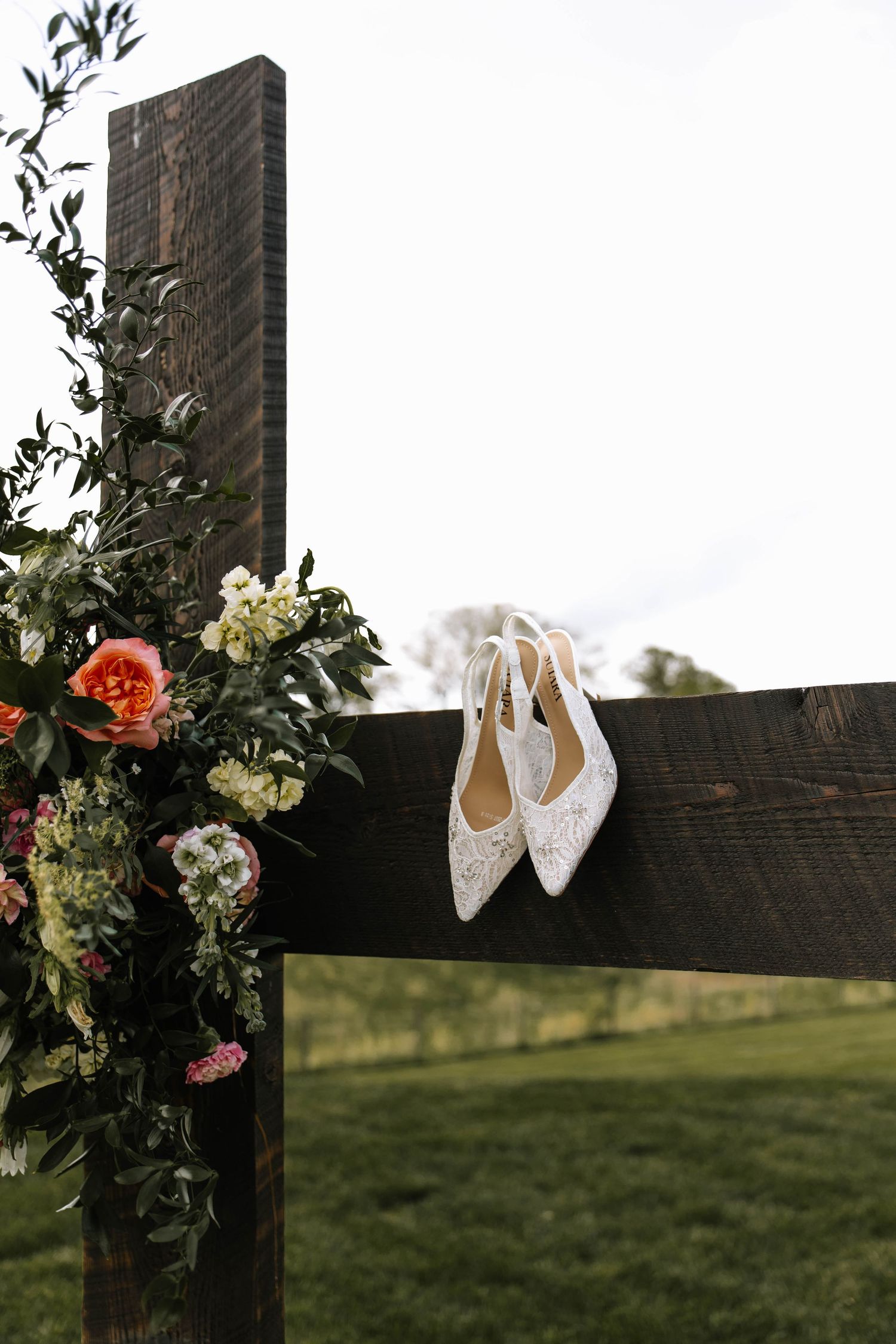 White lace bridal heels hanging on a rustic wooden arch adorned with colorful florals and greenery outdoors.