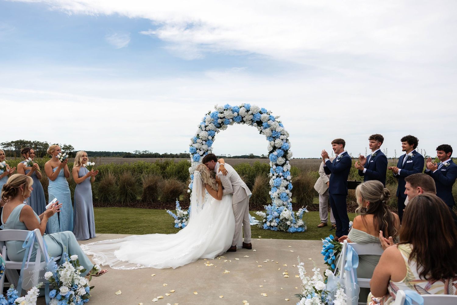 Couple kissing under a blue floral arch at an outdoor wedding ceremony with guests in blue attire.