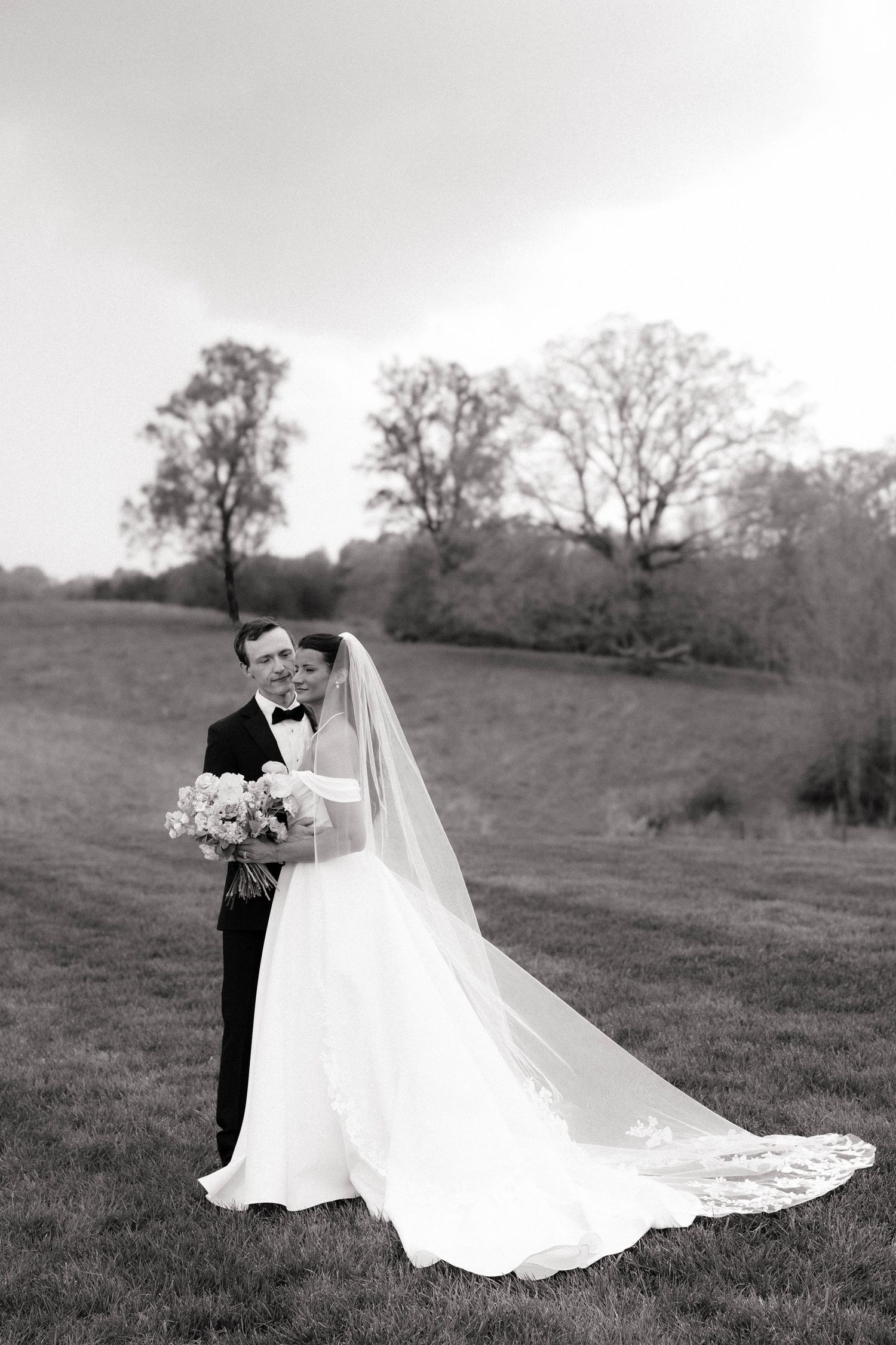 Black and white wedding photo of bride in flowing veil and groom in tuxedo embracing in a lush green field.