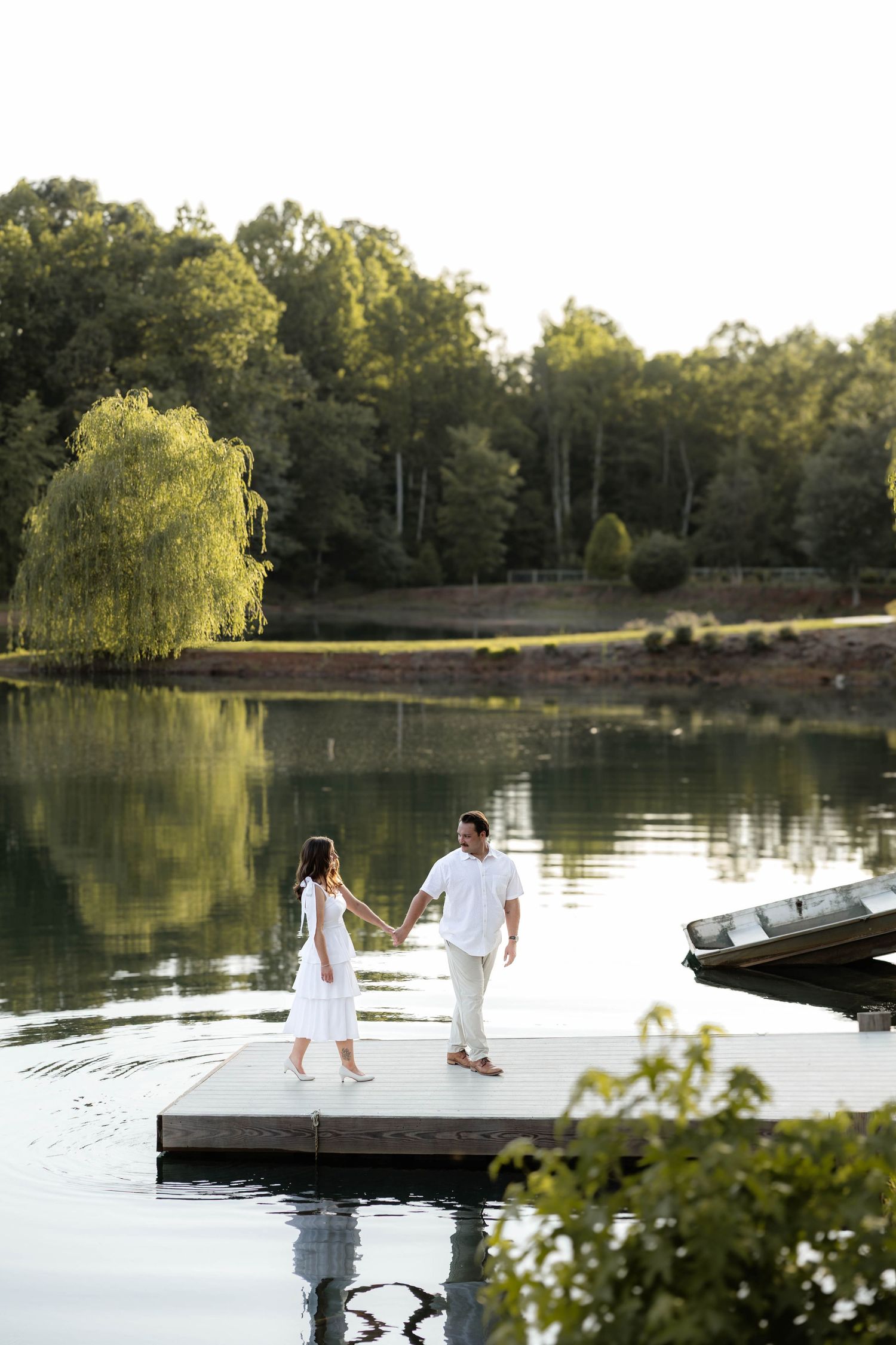 Couple in white outfits holding hands on a lakeside dock surrounded by lush green trees and calm reflective water.