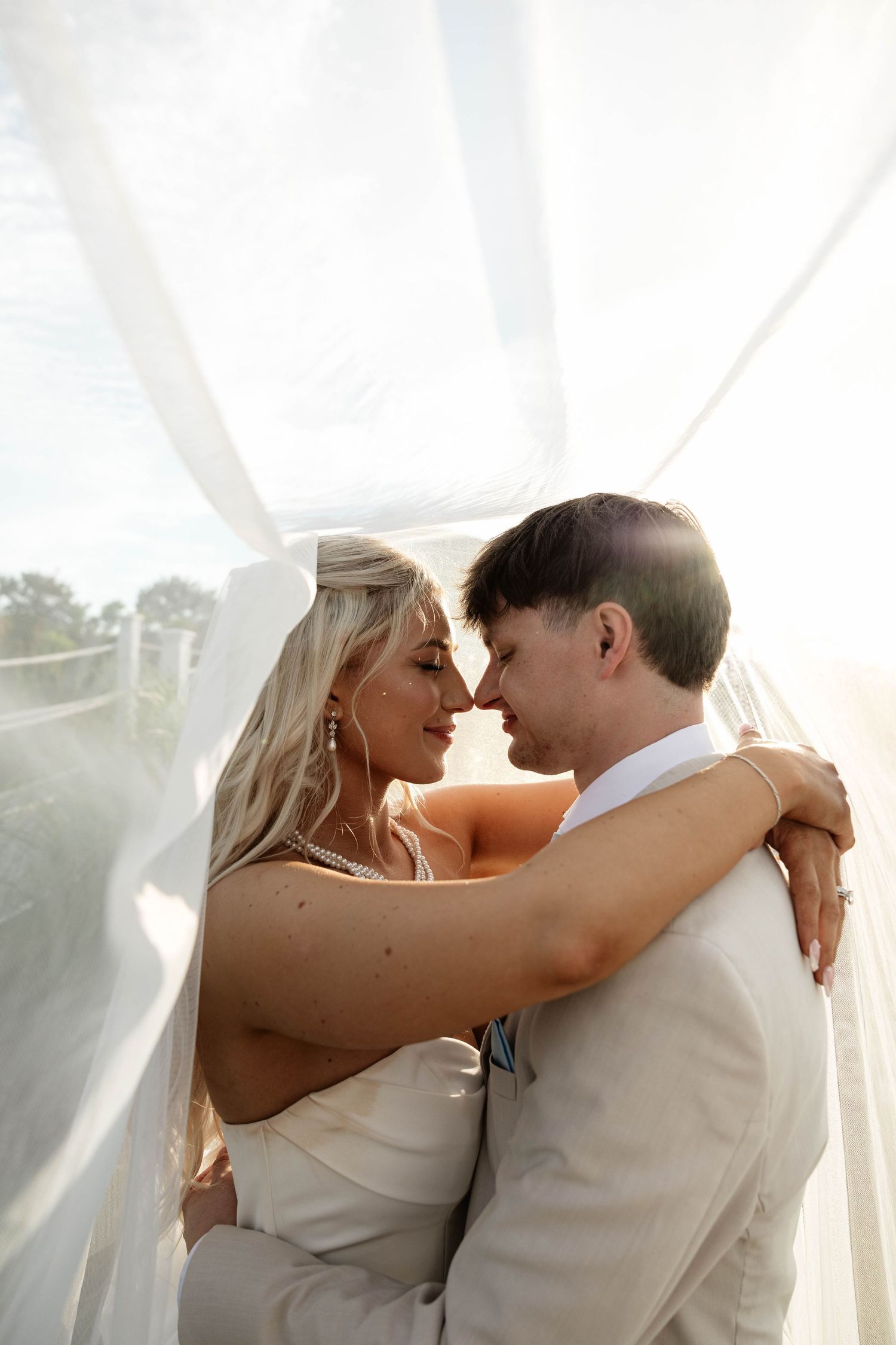 Bride and groom embrace under flowing white veil, sharing an intimate moment in soft backlit light.
