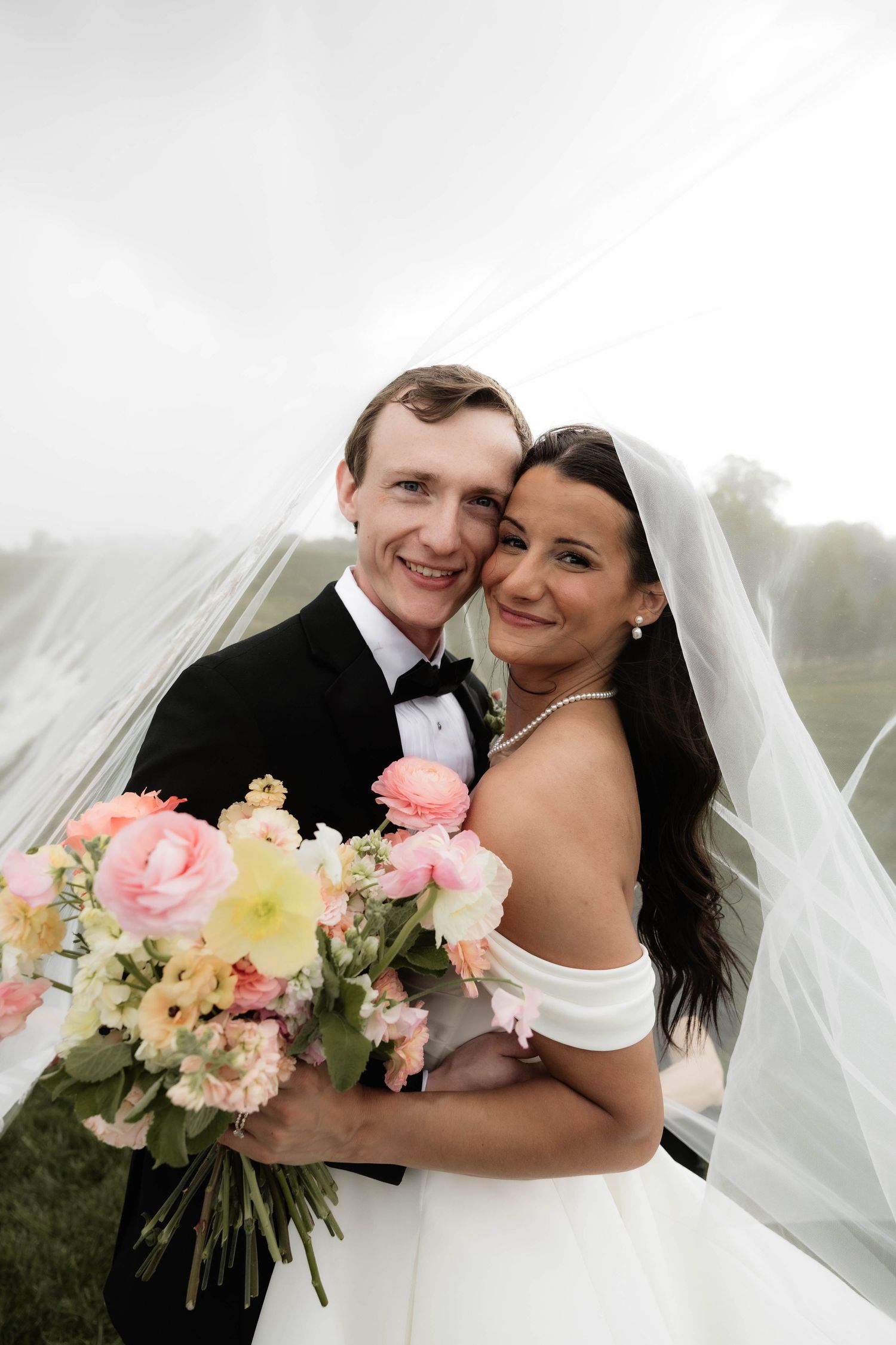 A smiling bride and groom pose under a flowing veil, holding a pink and yellow bouquet on their wedding day.