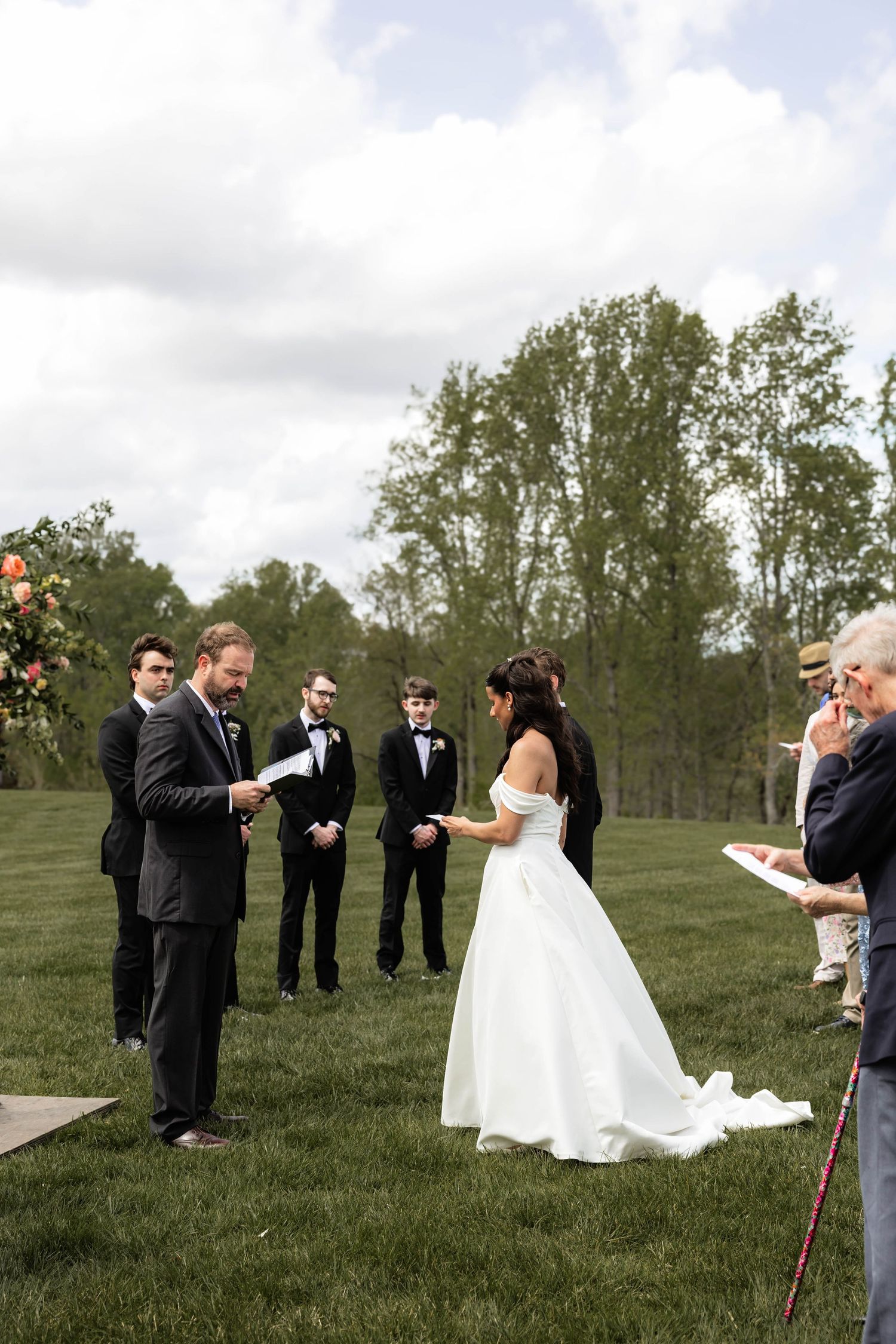 Outdoor wedding ceremony on green lawn with bride in white gown and groom reading vows, groomsmen and guests in background.
