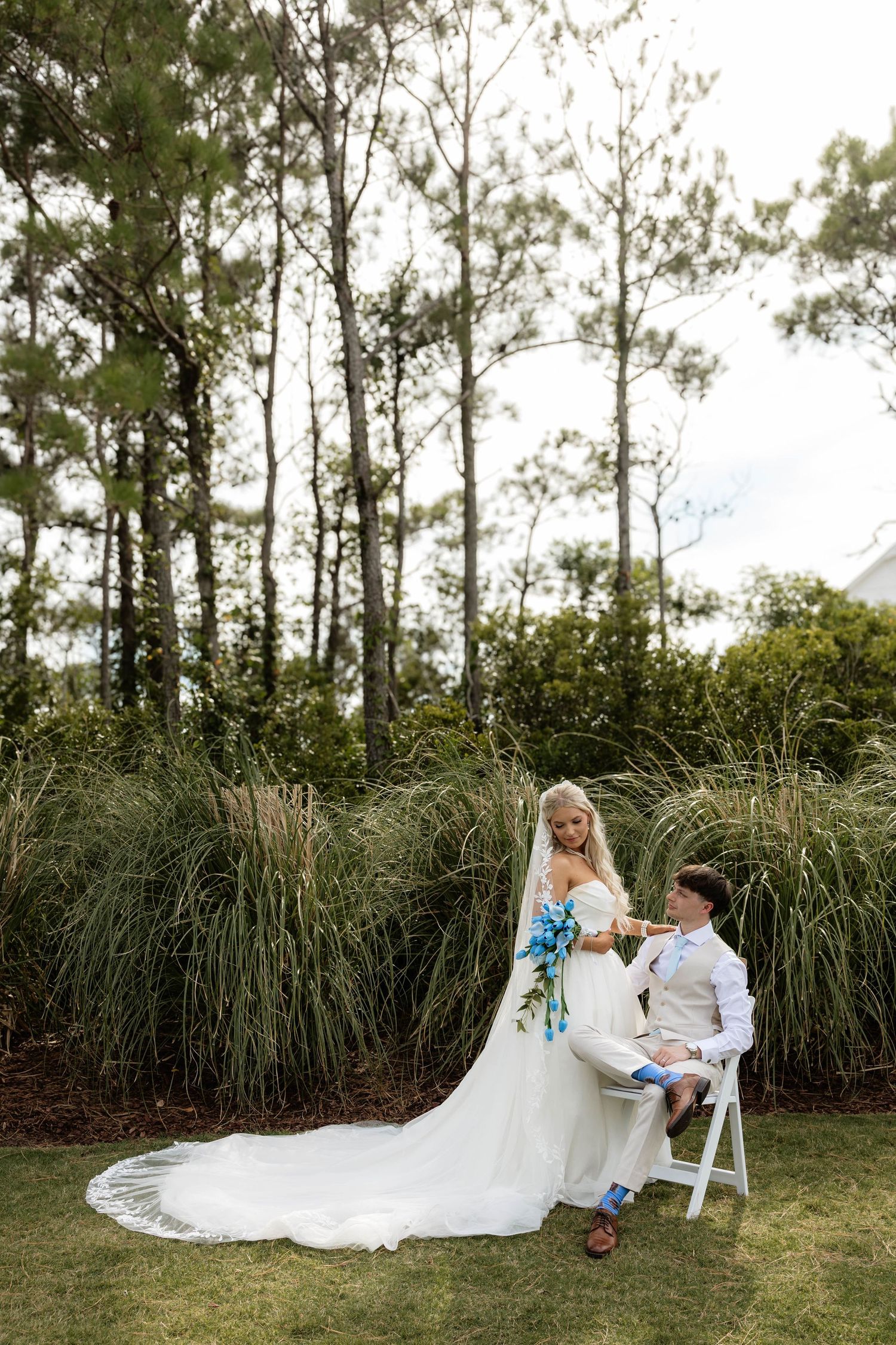 Bride in white gown with long veil holds blue bouquet beside groom seated in white chair on lush garden lawn.