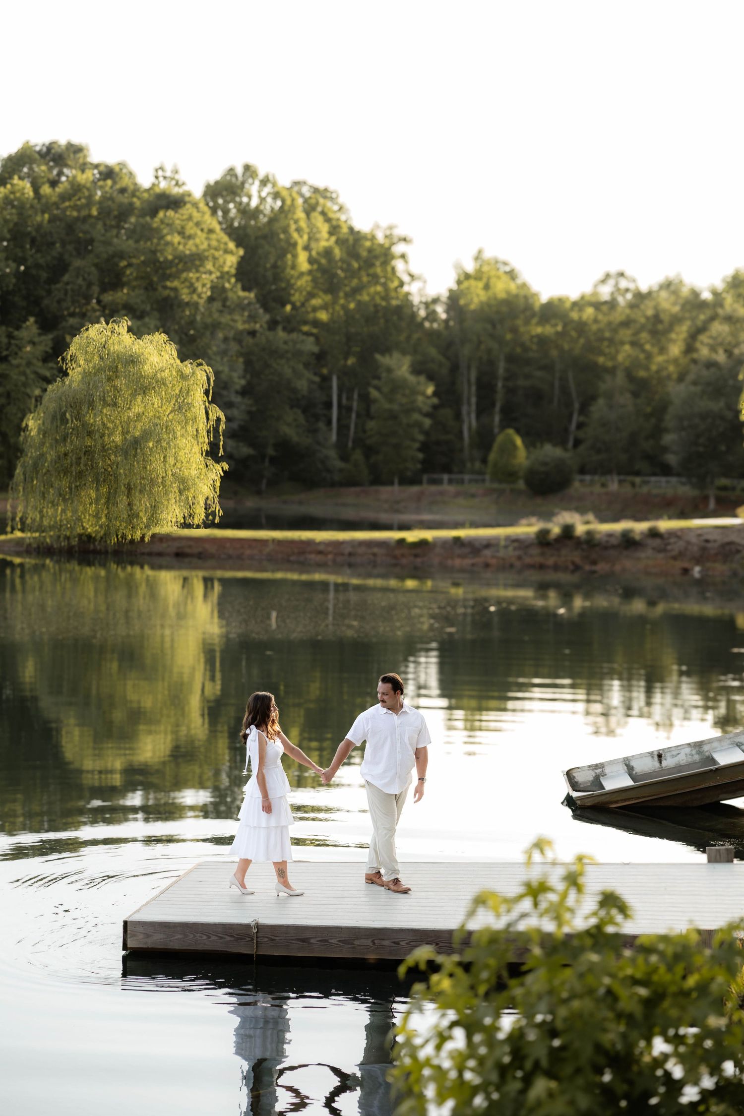 Couple in white holding hands on a wooden dock by a serene lake surrounded by lush green trees at golden hour.