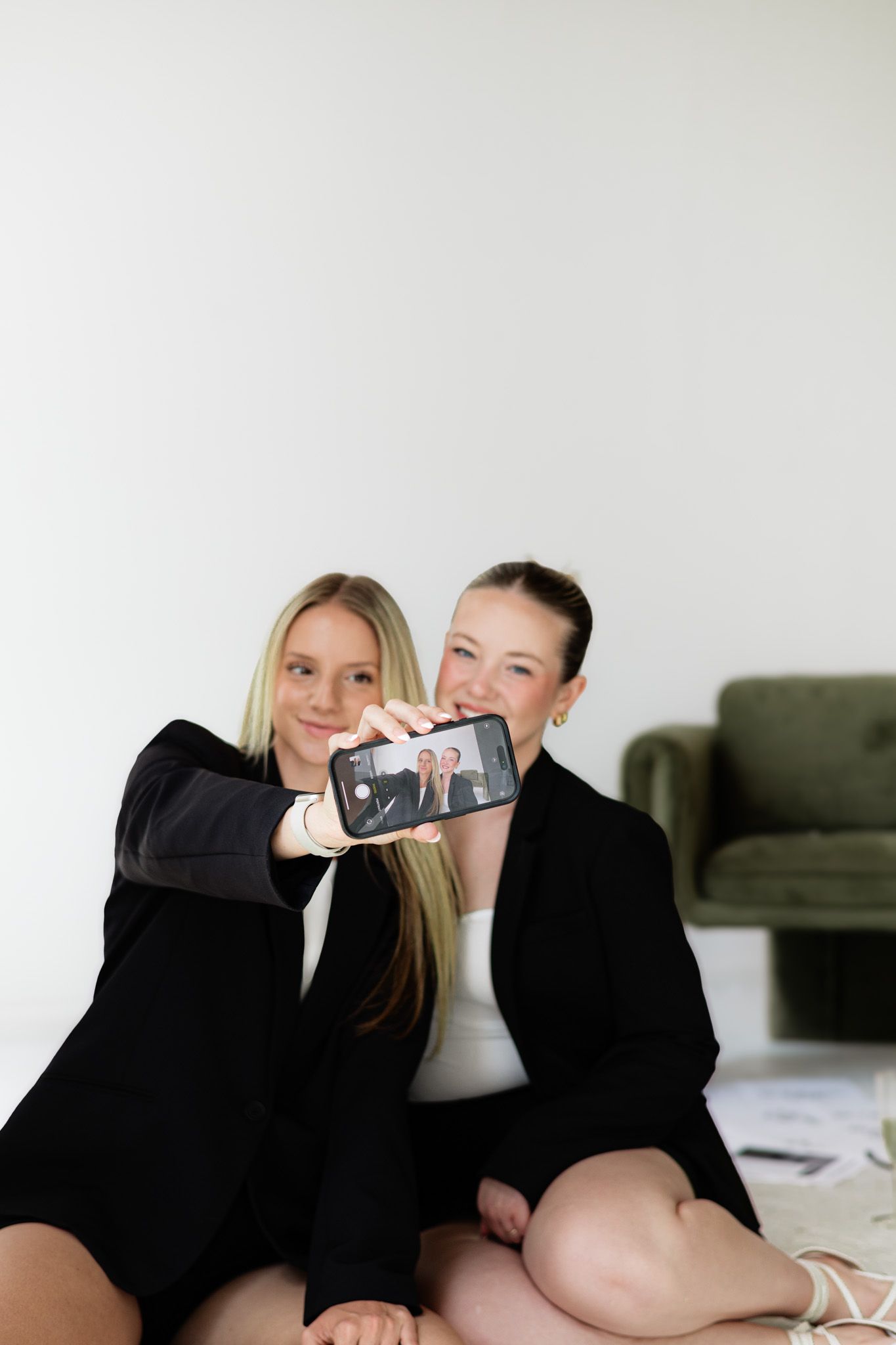 Two young women in black blazers sitting on floor, smiling while holding up a smartphone showing their selfie photo.
