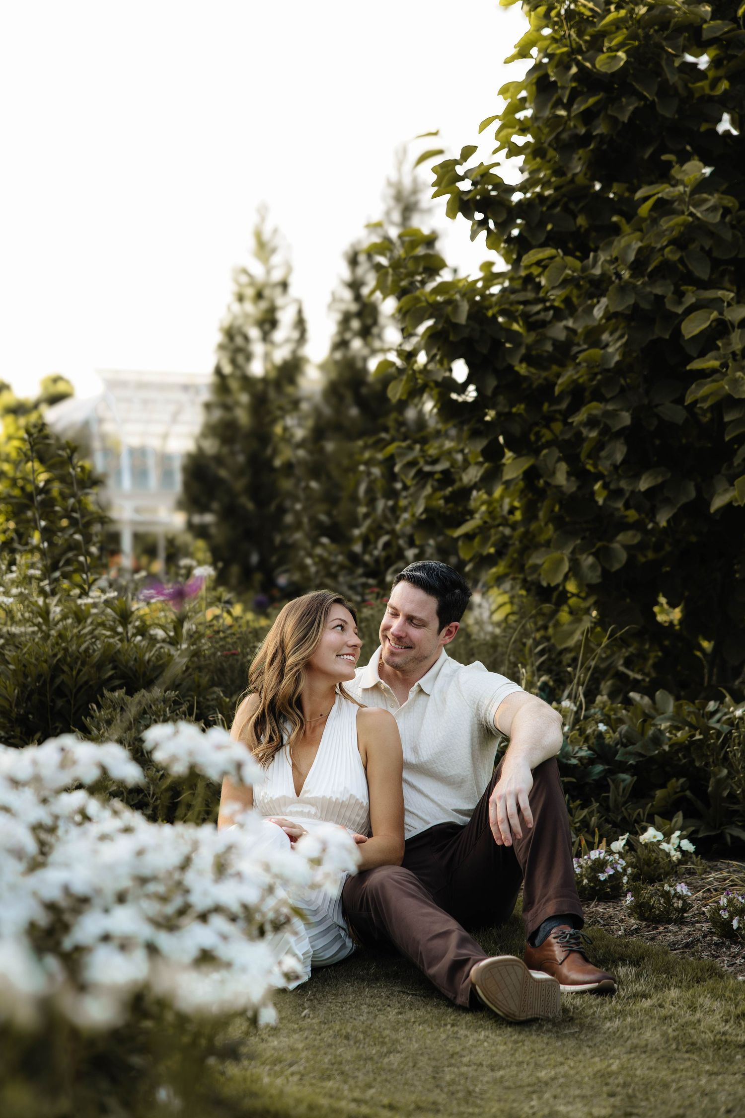 A smiling couple sits together on lush green grass surrounded by white flowers and tall trees in a garden.