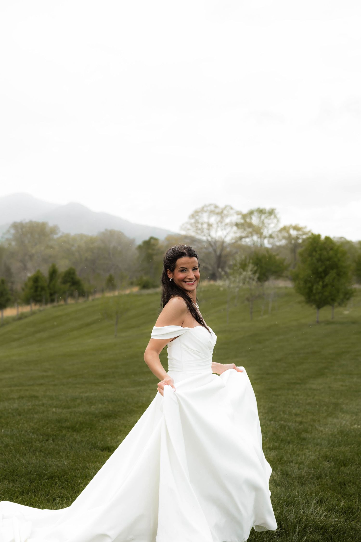 Smiling bride in off-shoulder white gown twirling on green lawn with mountain backdrop.